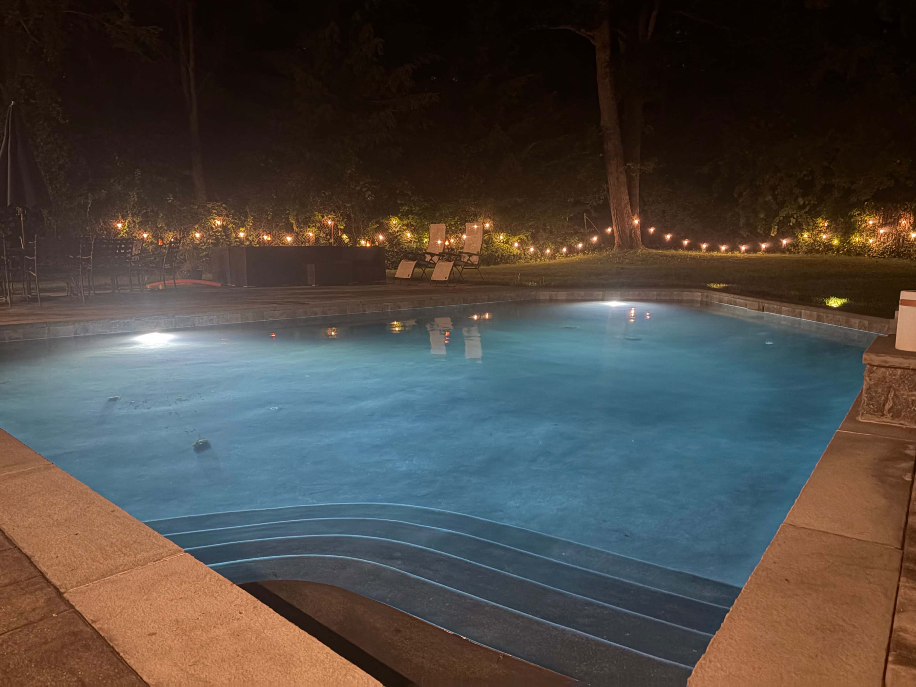 The image shows a dimly lit pool area surrounded by trees, with a pool illuminated by underwater lights and string lights hanging in the background.