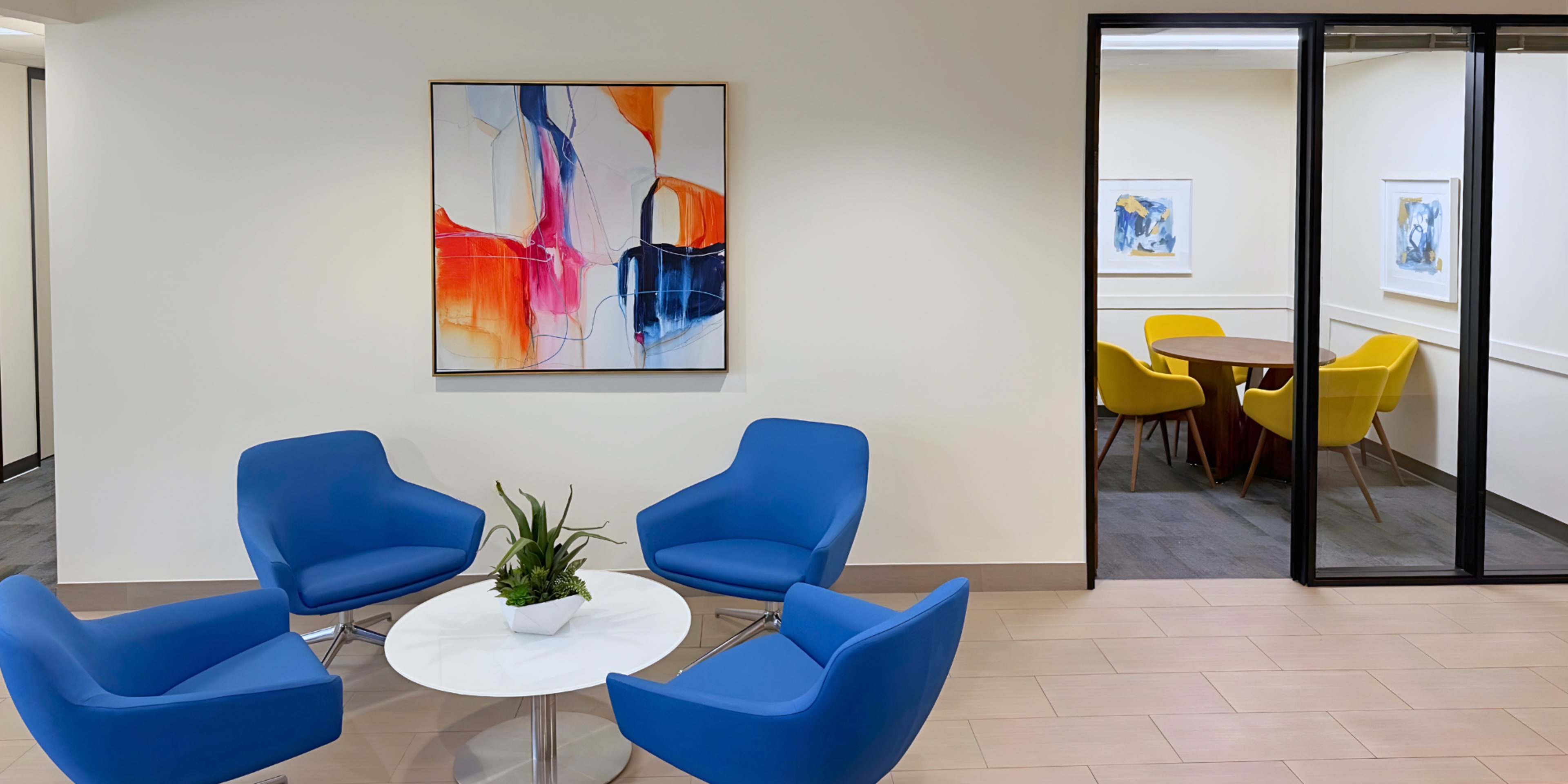 A seating area features blue chairs surrounding a round table, with a colorful abstract painting on the wall and a glass partition revealing another meeting room with yellow chairs.