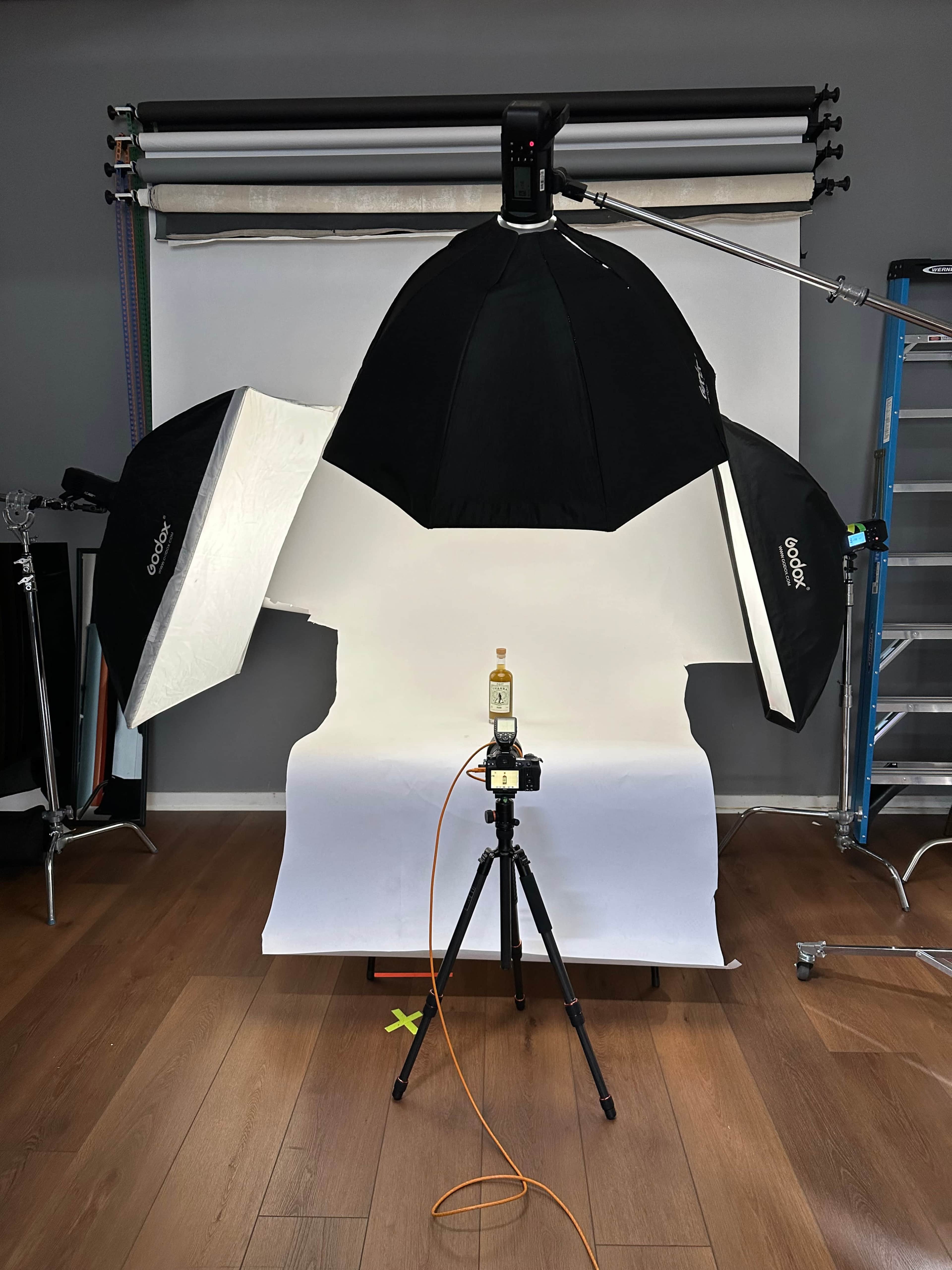 The image shows a photography setup with a camera on a tripod, an illuminated backdrop, and two large softboxes positioned to light a bottle placed on the ground.