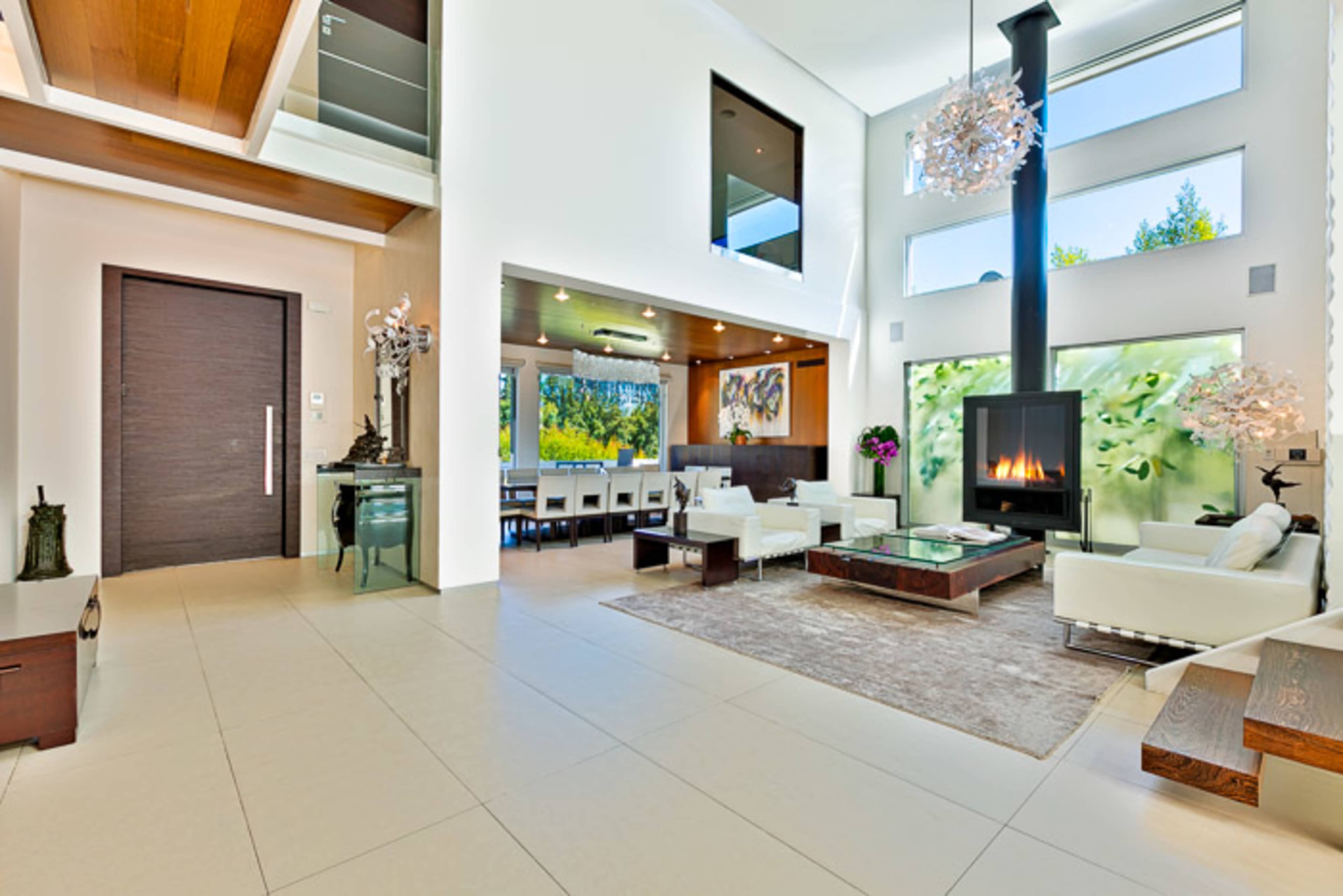 The image depicts a modern living room with high ceilings, large windows, and contemporary furniture arranged around a central fireplace.