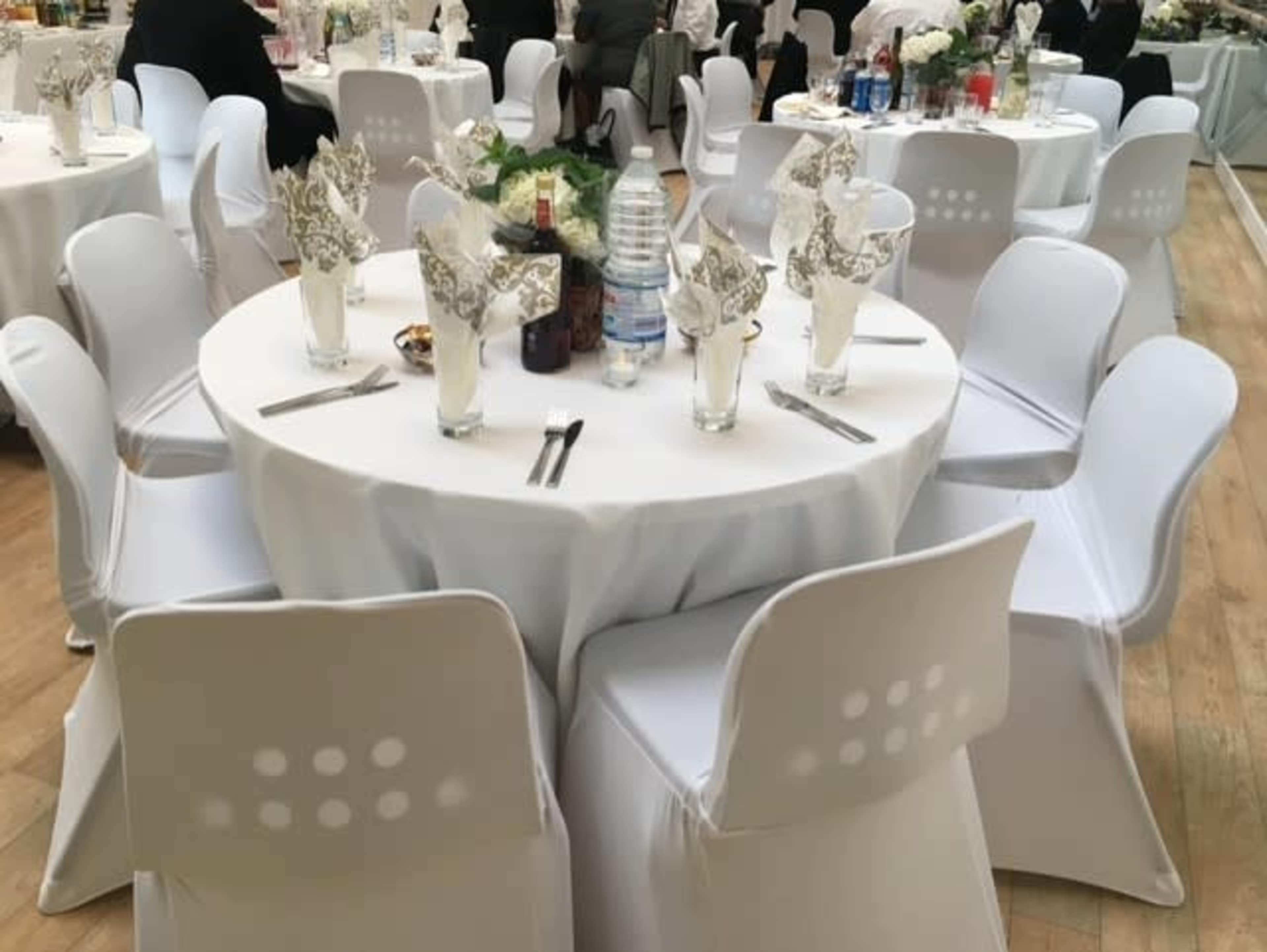 A round dining table is set with glasses, silverware, and folded napkins, surrounded by white chairs in a banquet hall.