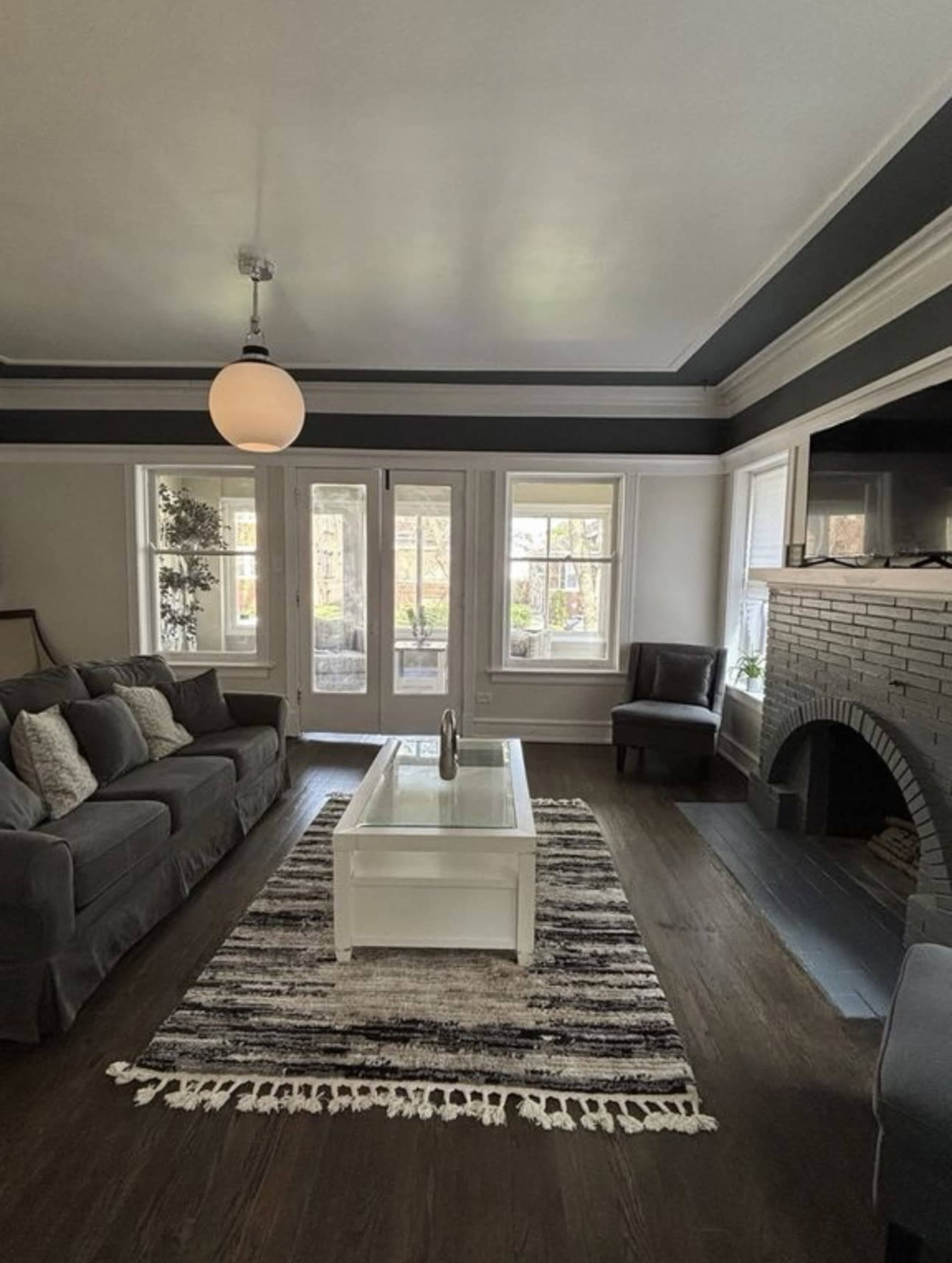 The room features a gray sofa and a white coffee table on a striped rug, with large windows allowing natural light to enter.