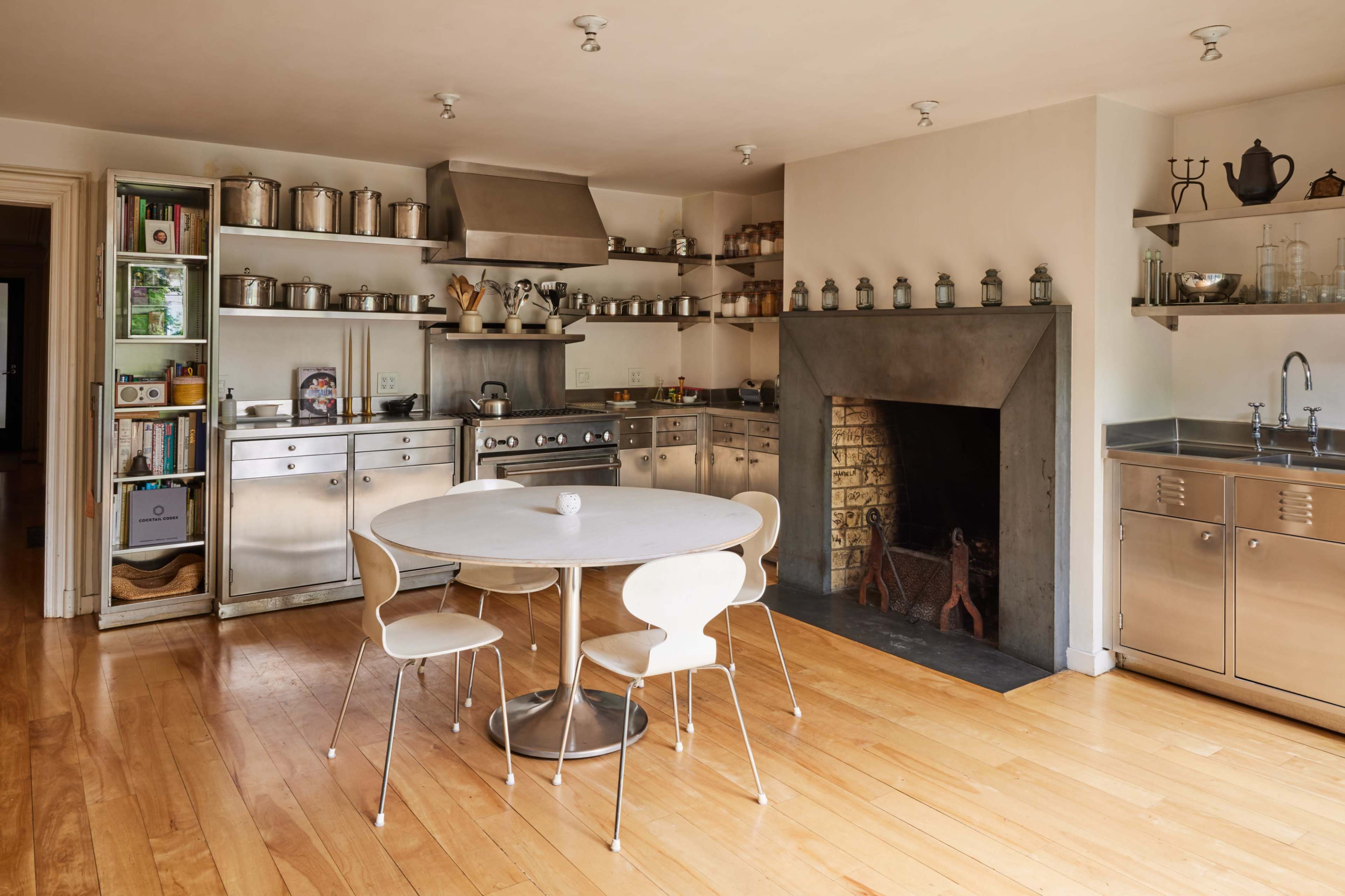 A modern kitchen with stainless steel appliances, shelves filled with jars, and a round table surrounded by four white chairs.