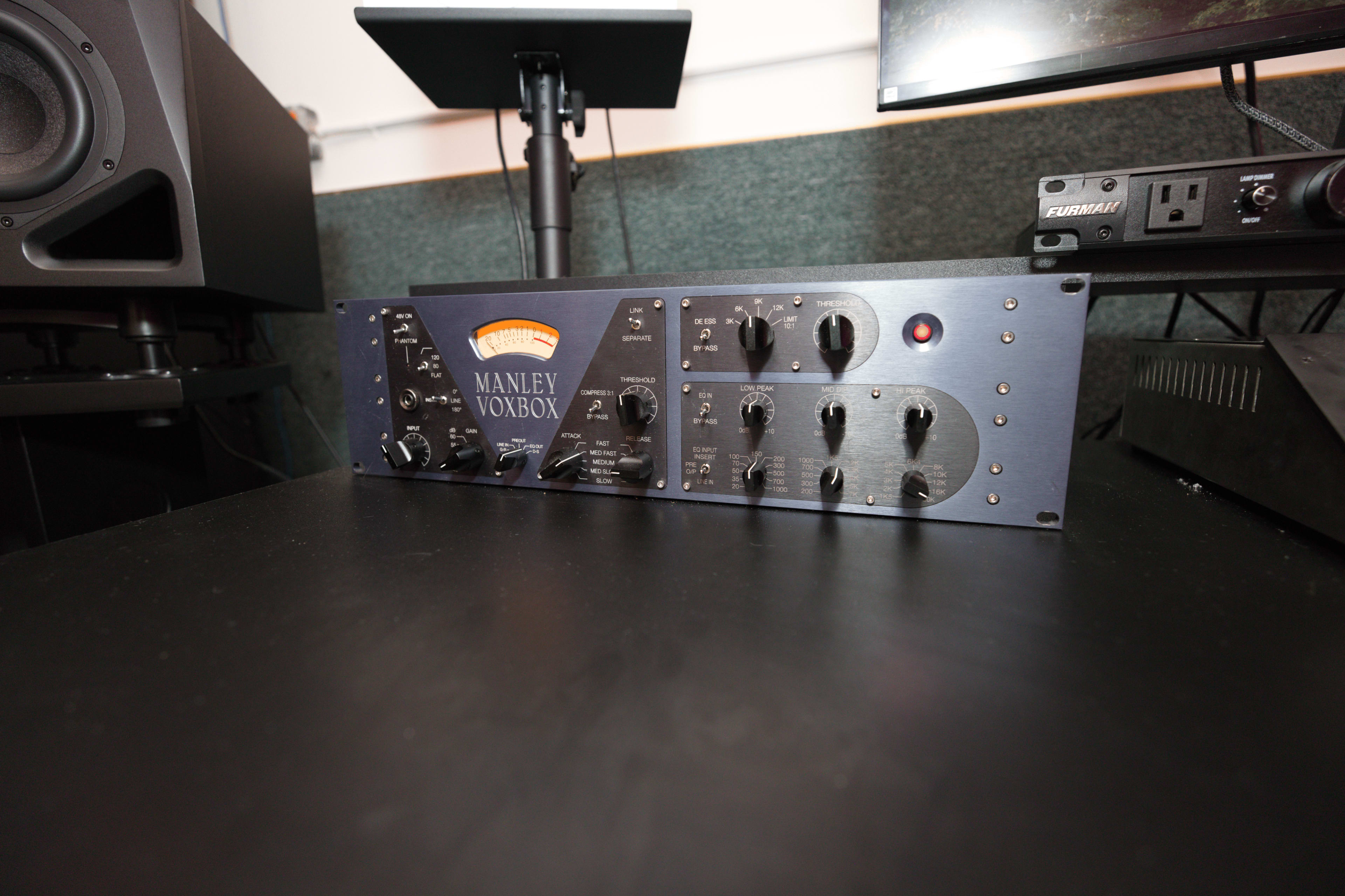 The image shows a Manley Voxbox, a piece of audio equipment, placed on a black desk with studio monitors and a computer monitor in the background.