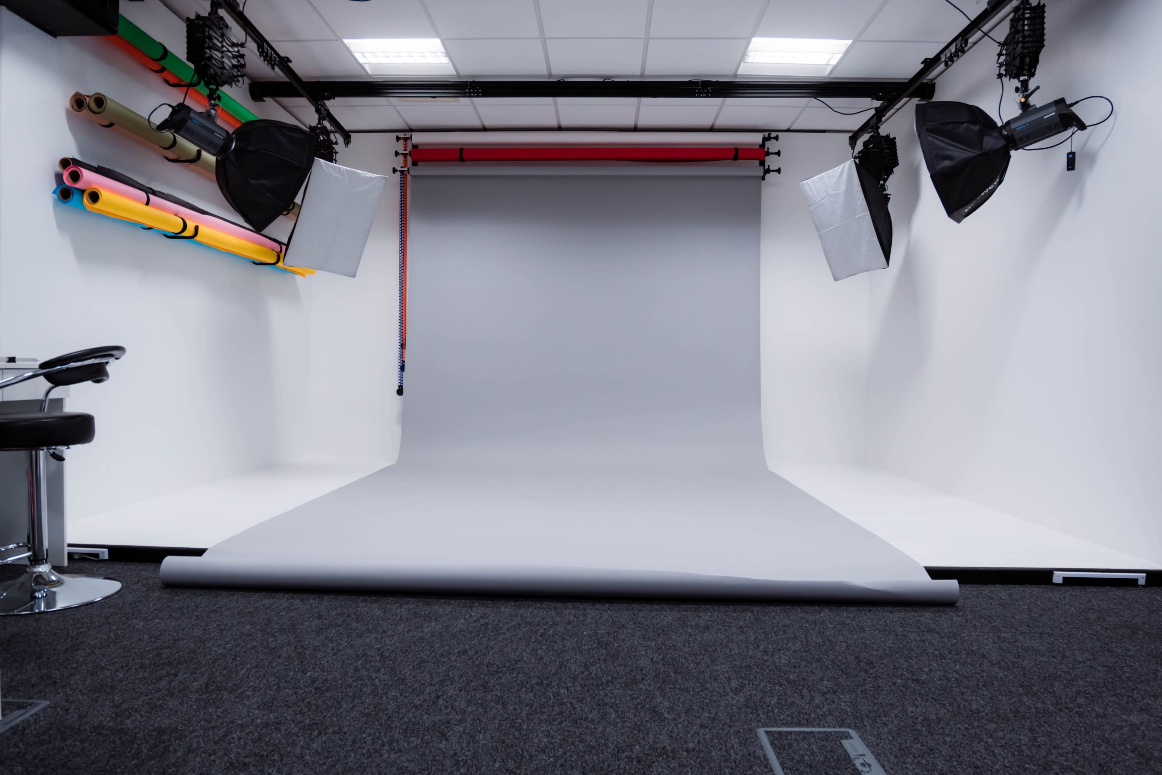 A photography studio with a gray backdrop, two softbox lights, and colorful rolls of backdrop paper mounted on the wall.