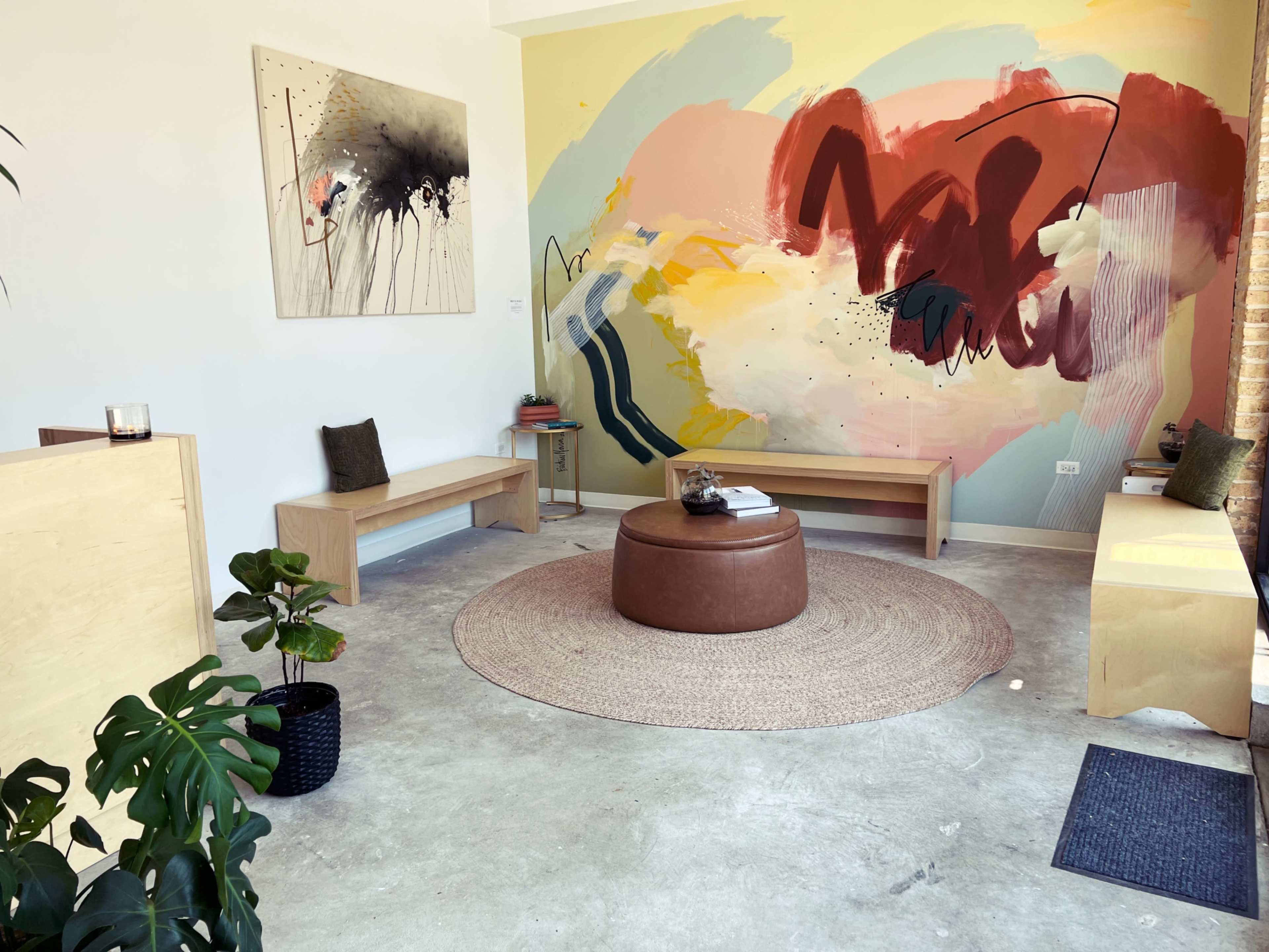 The image shows a modern interior space featuring a colorful mural, wooden benches, a round brown ottoman, and potted plants.