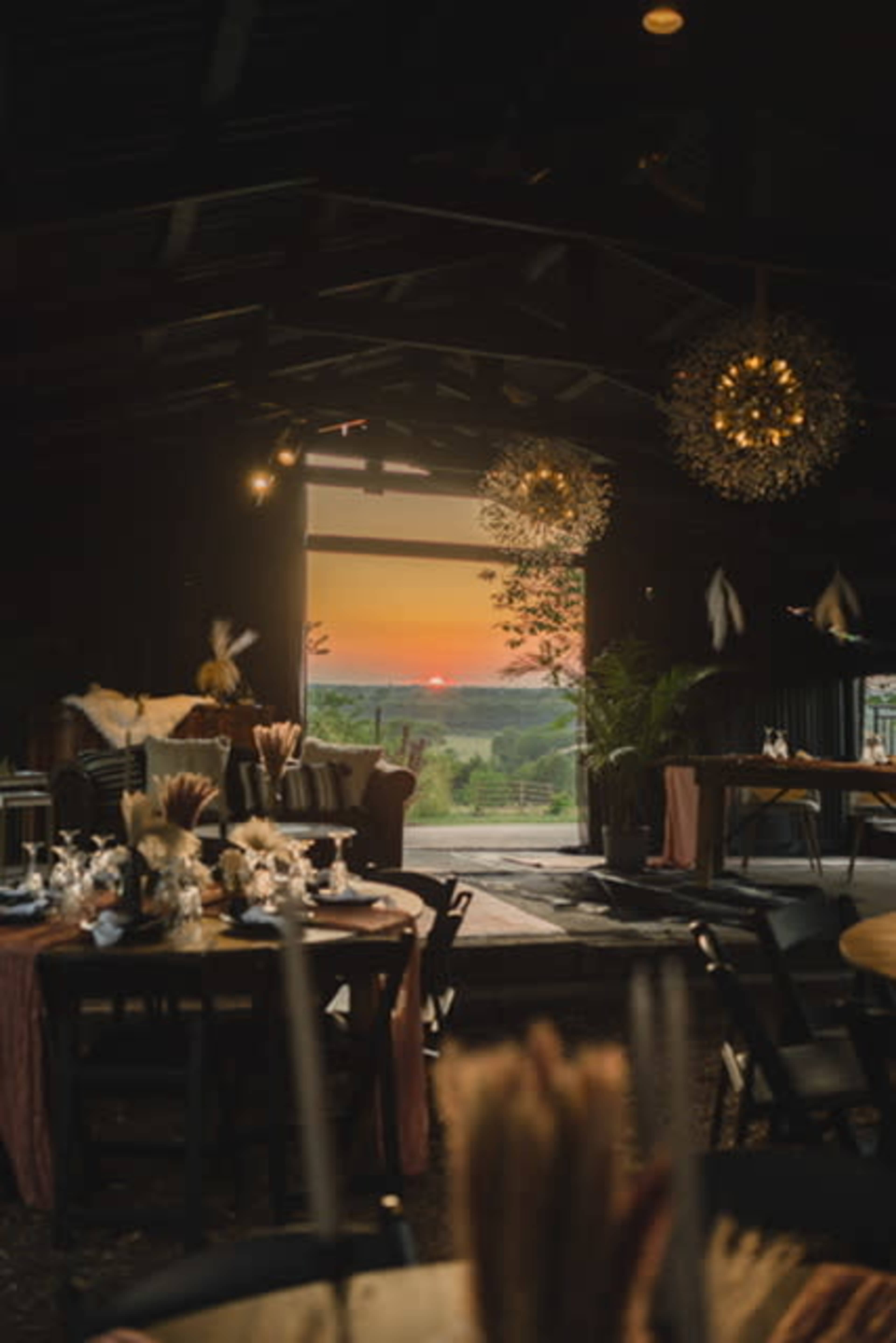 A rustic dining room features elegantly set tables with floral arrangements, while the sun sets beyond an open door, casting warm light into the space.