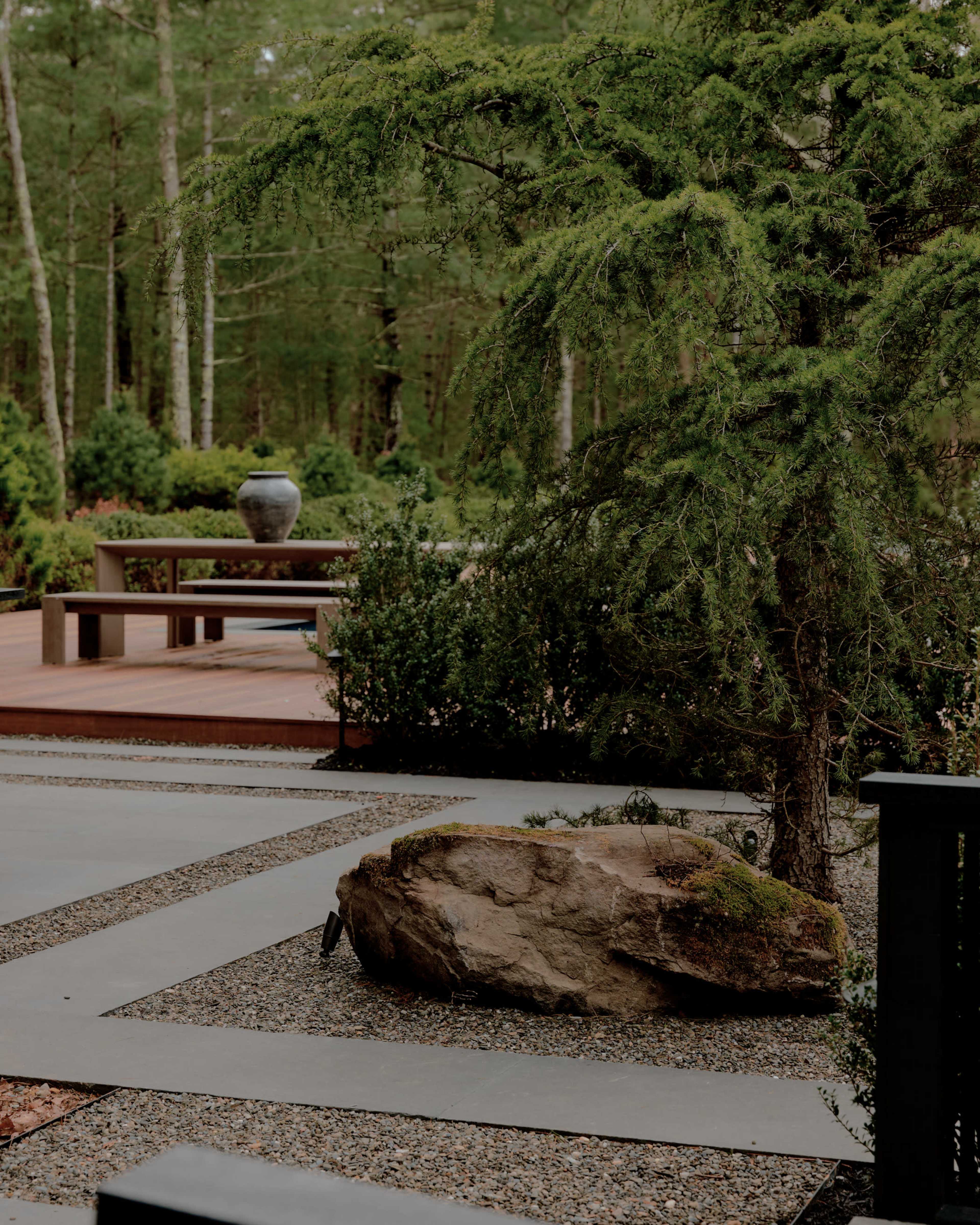 A stone sits amidst gravel pathways and greenery in a landscaped garden setting with a patio in the background.