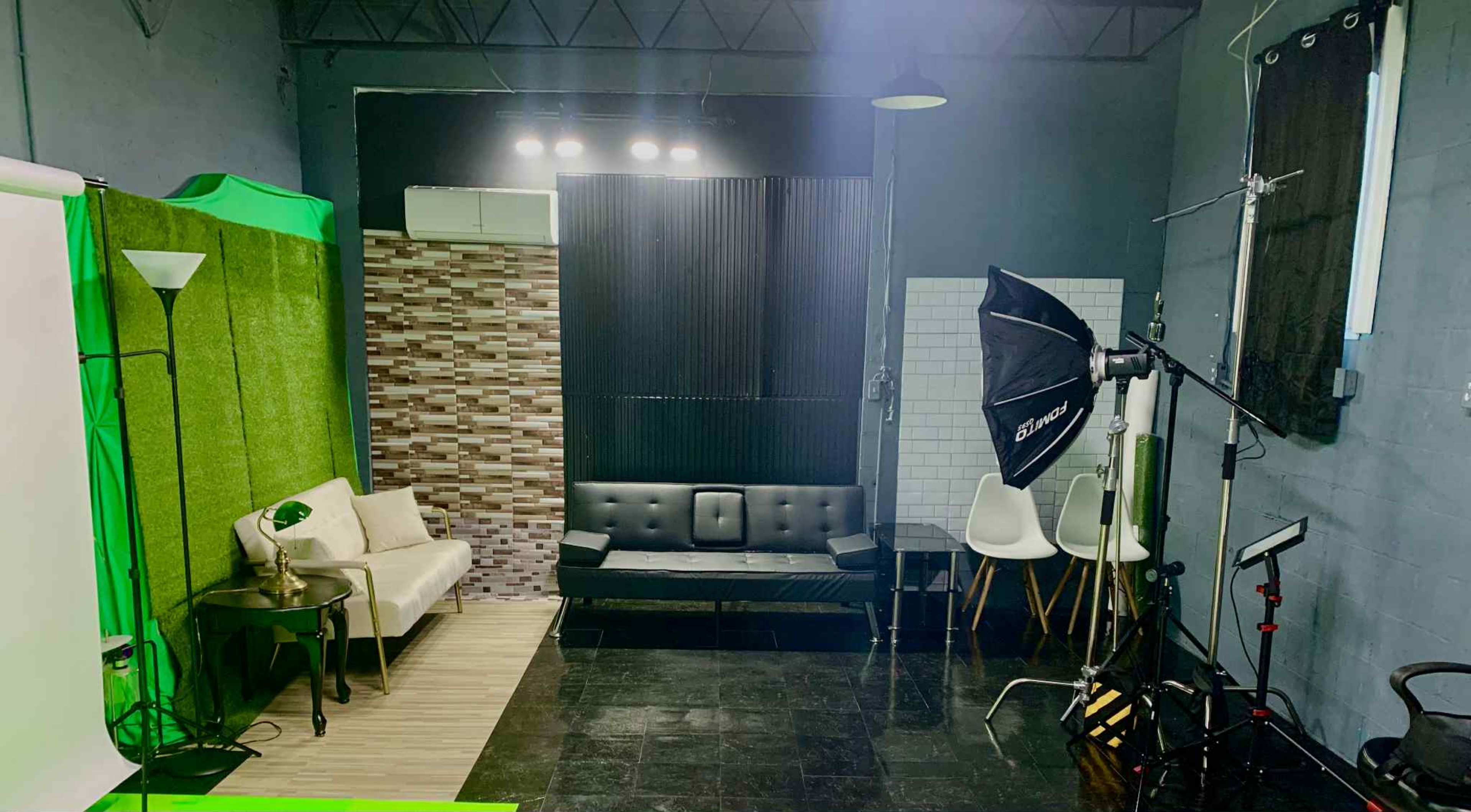The image shows a photo studio setup with a green screen area, a sofa, chairs, lighting equipment, and a textured wall.