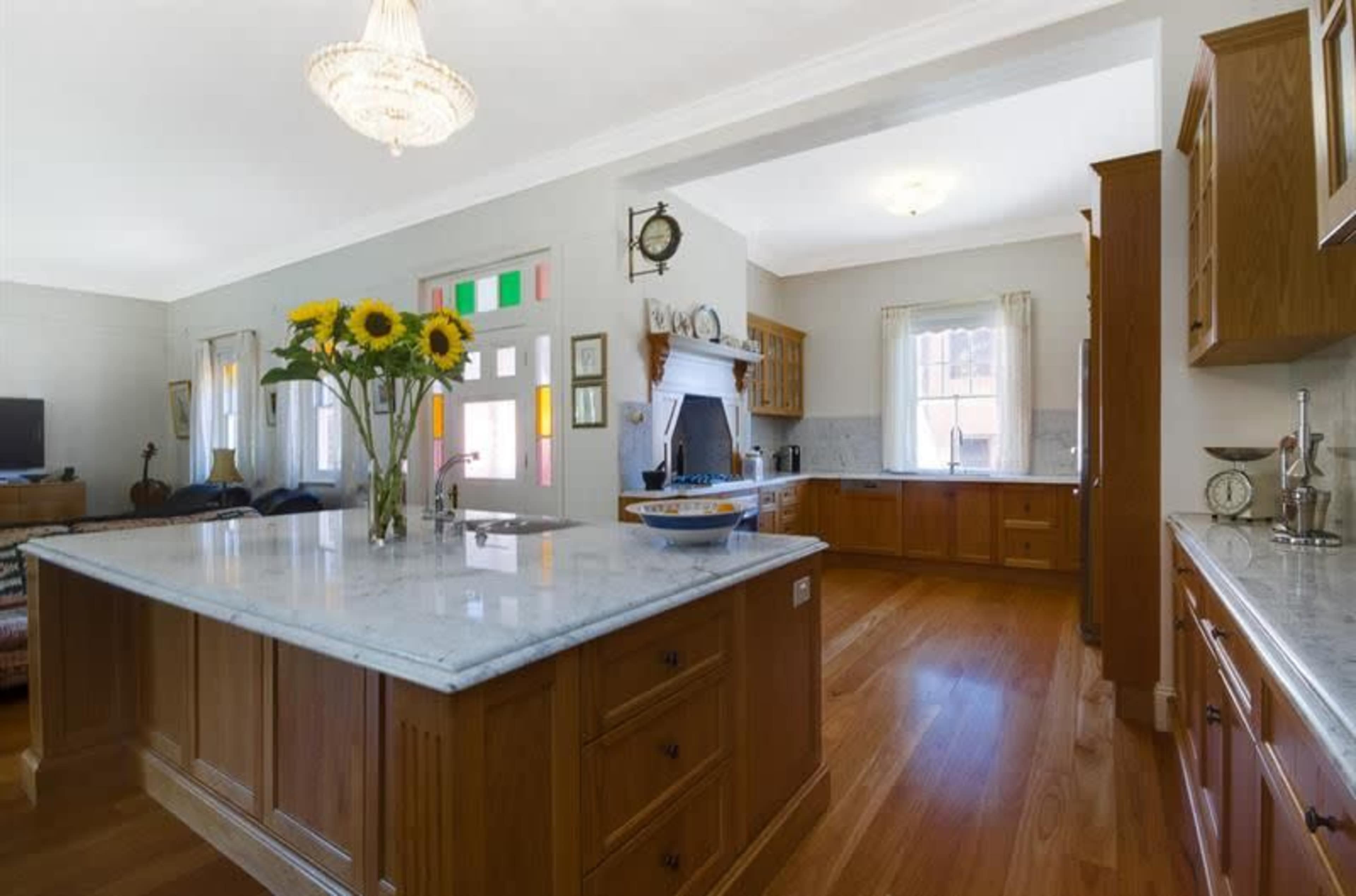 The image shows a bright kitchen with wooden cabinetry, marble countertops, and a vase of sunflowers on an island counter.