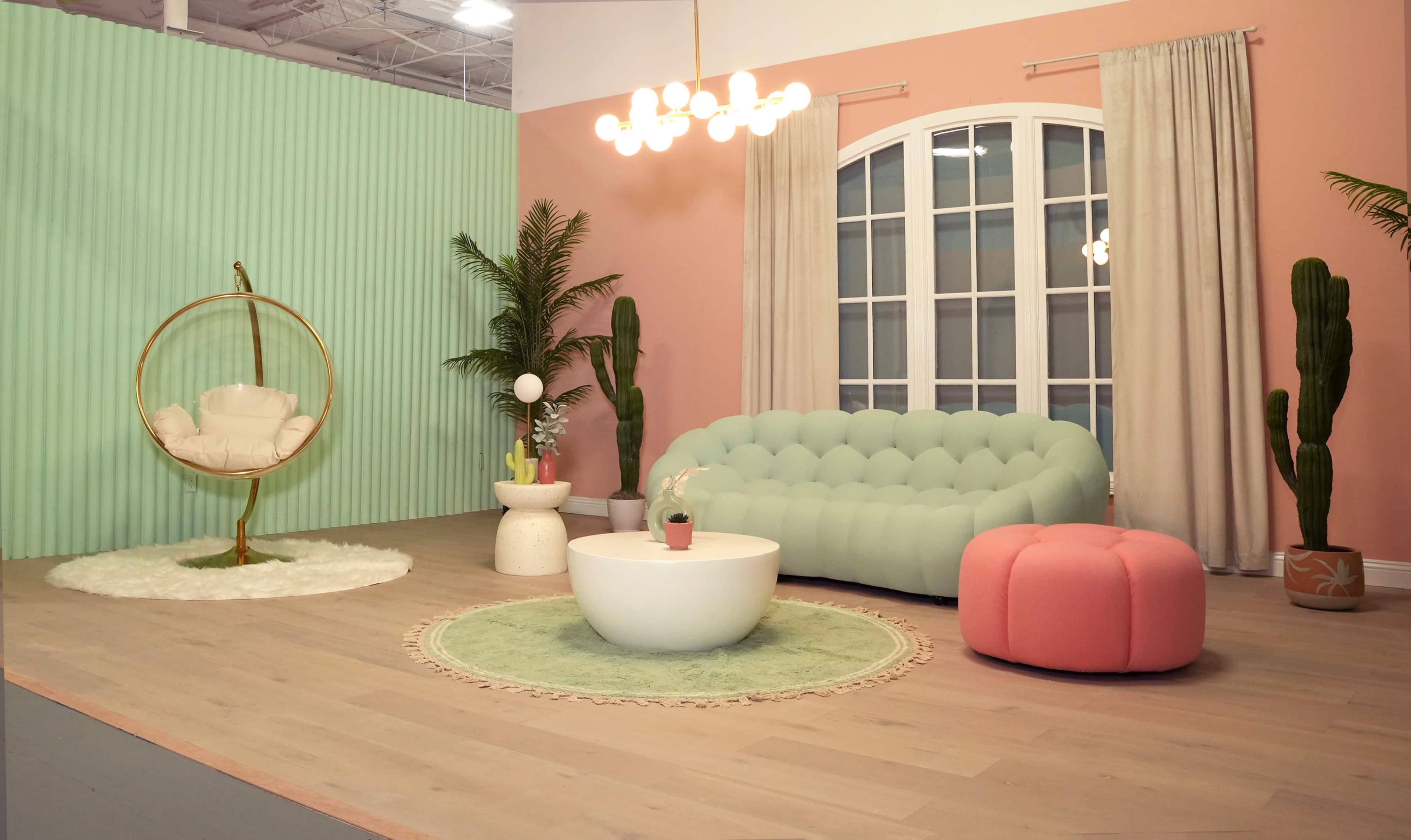 A brightly colored interior living space featuring a mint green couch, a circular white coffee table, a pink ottoman, and various decorative plants.