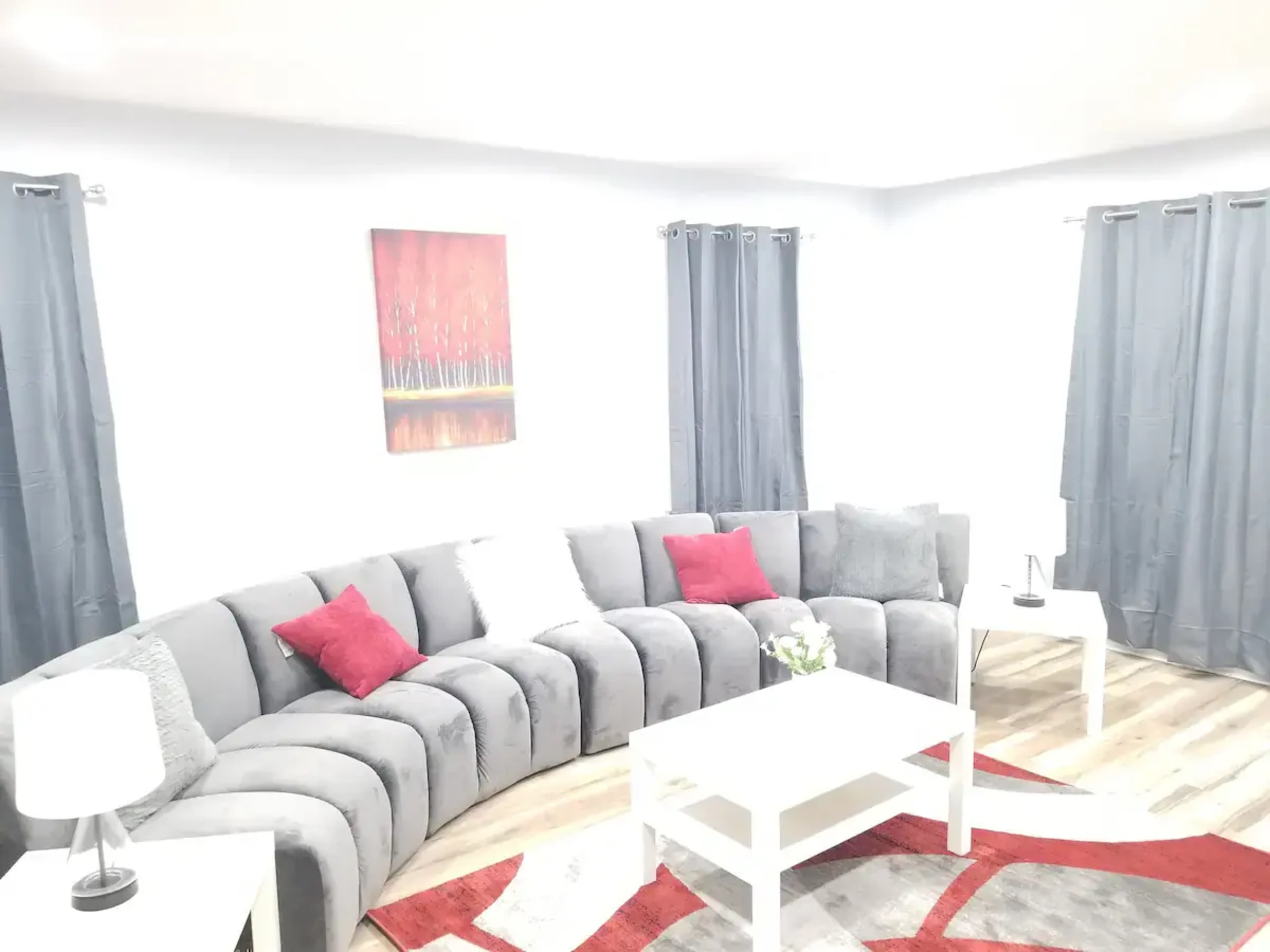 A modern living room with a curved gray sofa, a white coffee table, and red accent pillows, complemented by curtains and artwork on the walls.