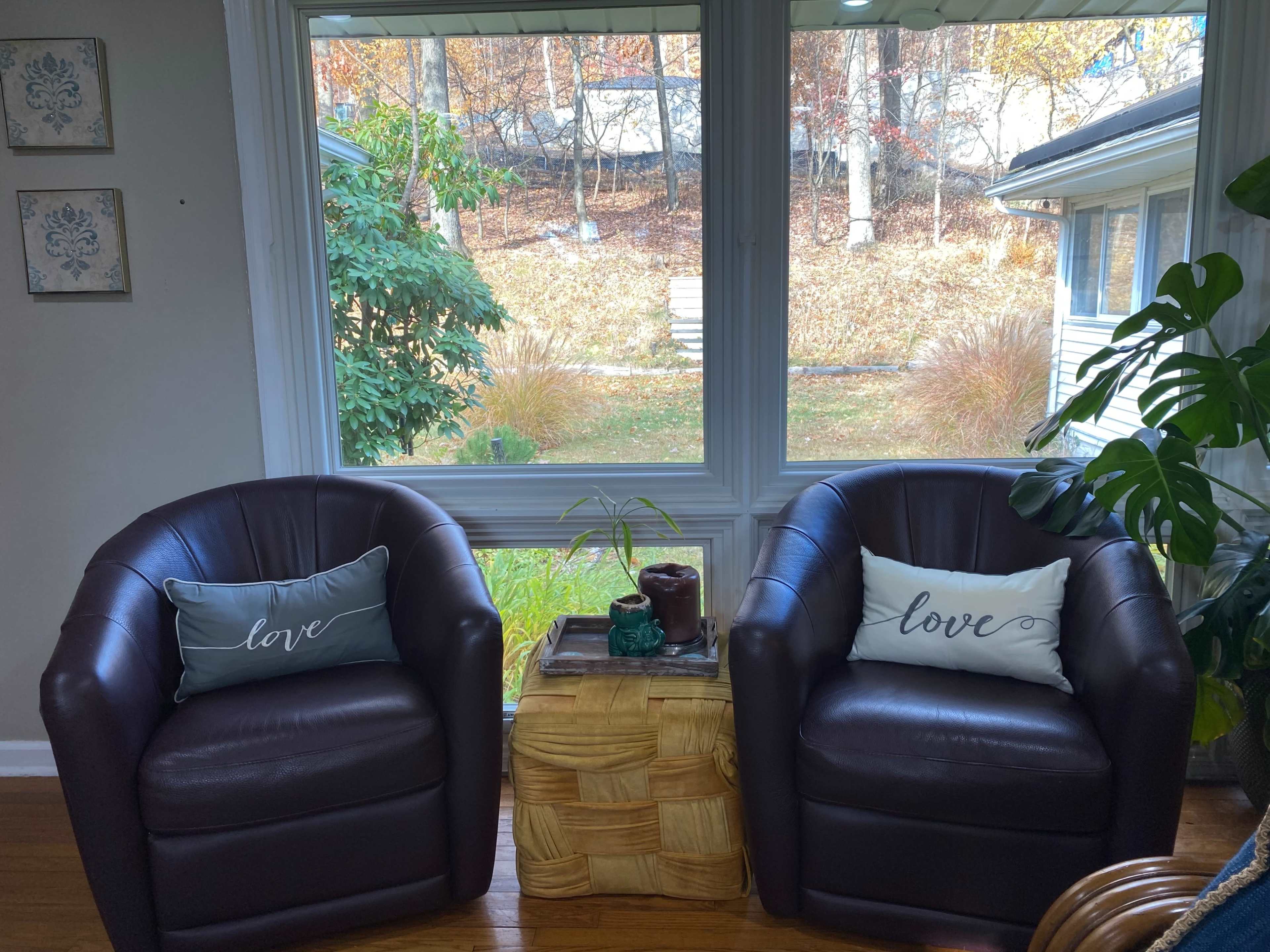 Two brown armchairs with decorative pillows sit across from each other in front of a large window overlooking a landscaped yard with trees.