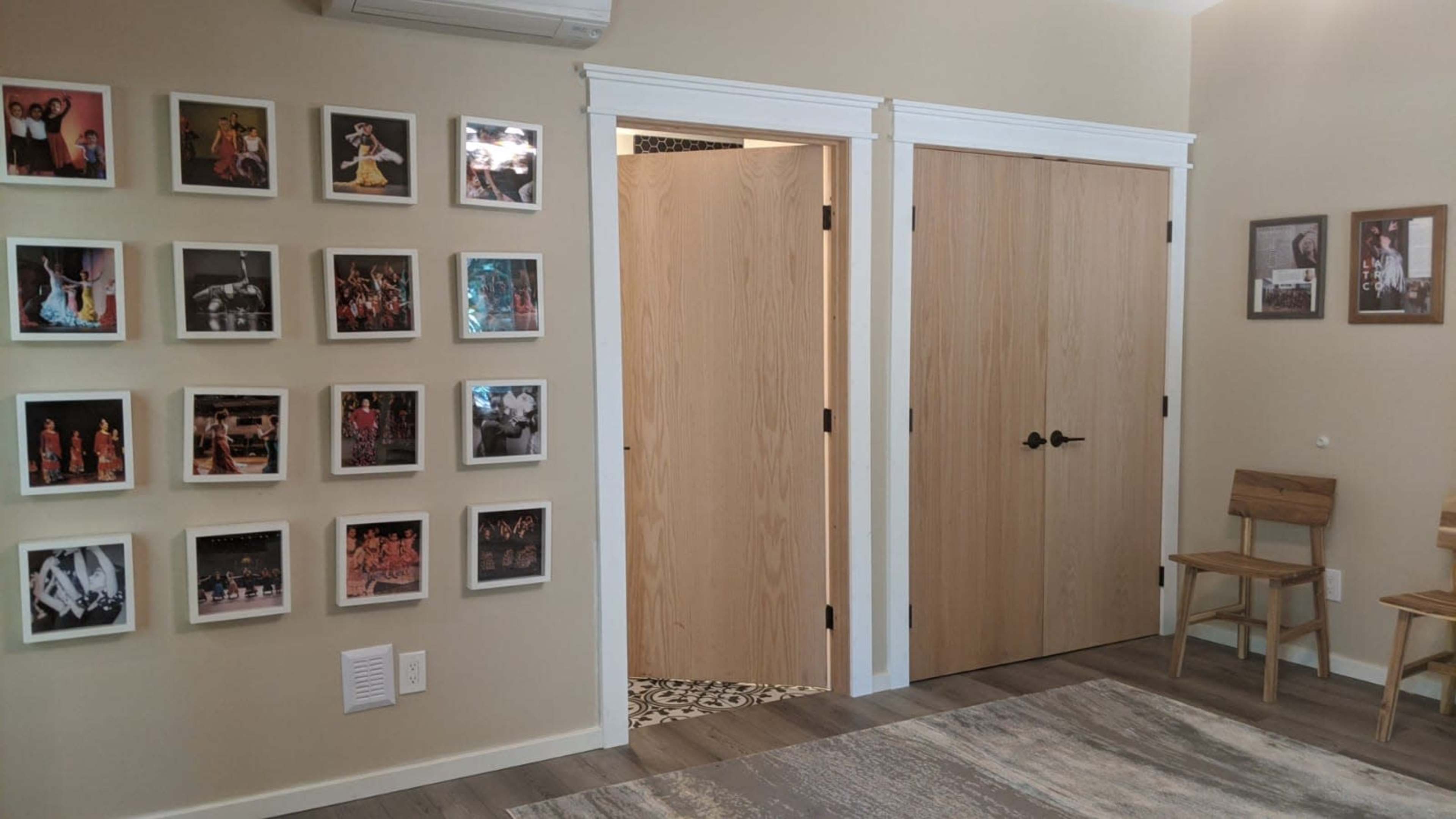 A room with a wall featuring a grid of framed photos, two wooden doors opposite each other, and two wooden chairs positioned nearby.