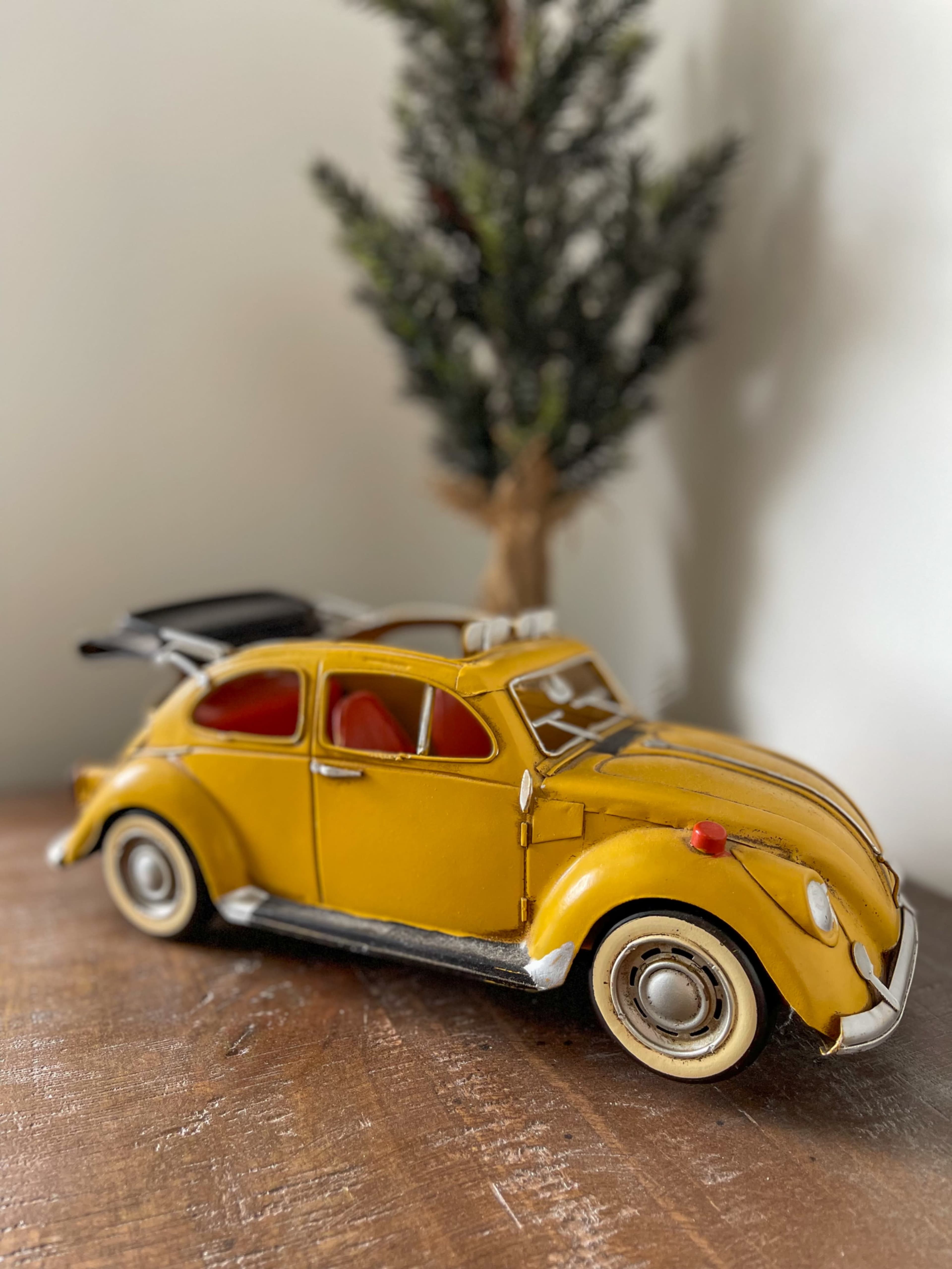 A vintage yellow toy car is positioned on a wooden surface beside a small evergreen plant.