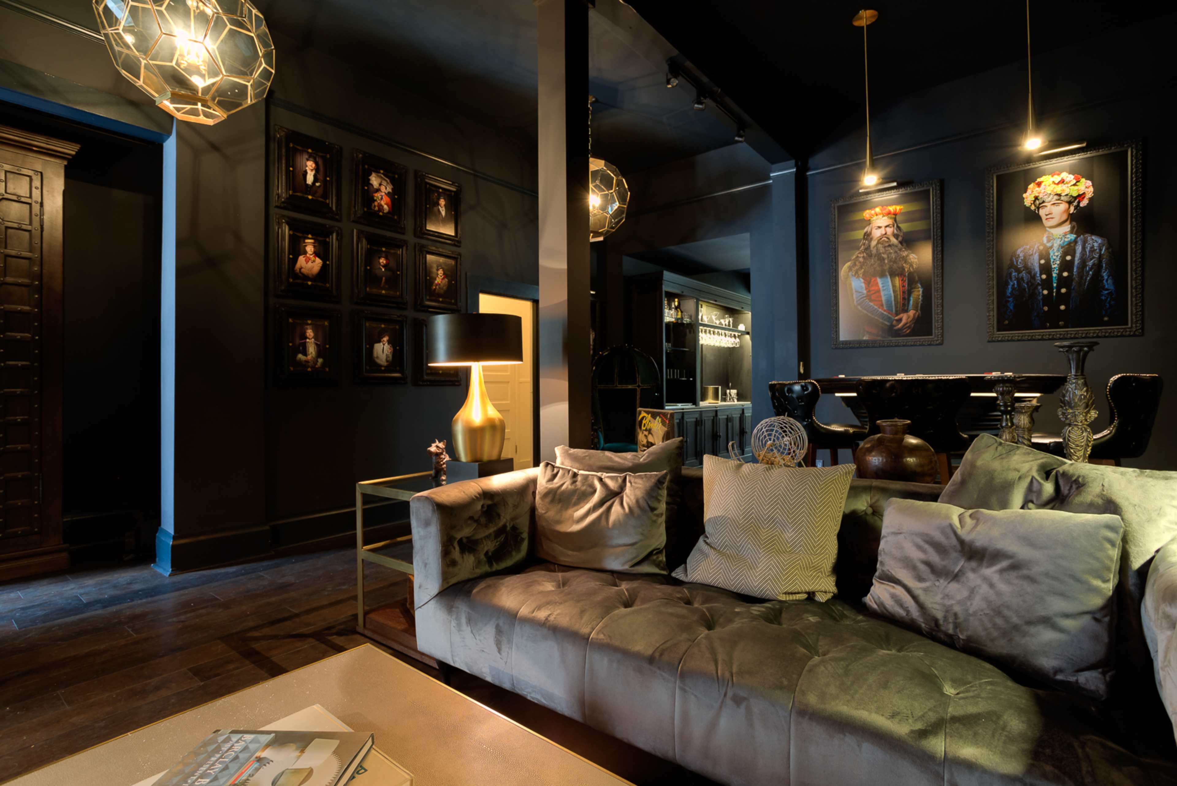 A stylish interior featuring a dark room with plush seating, modern lighting fixtures, framed portraits on the walls, and a sleek coffee table.
