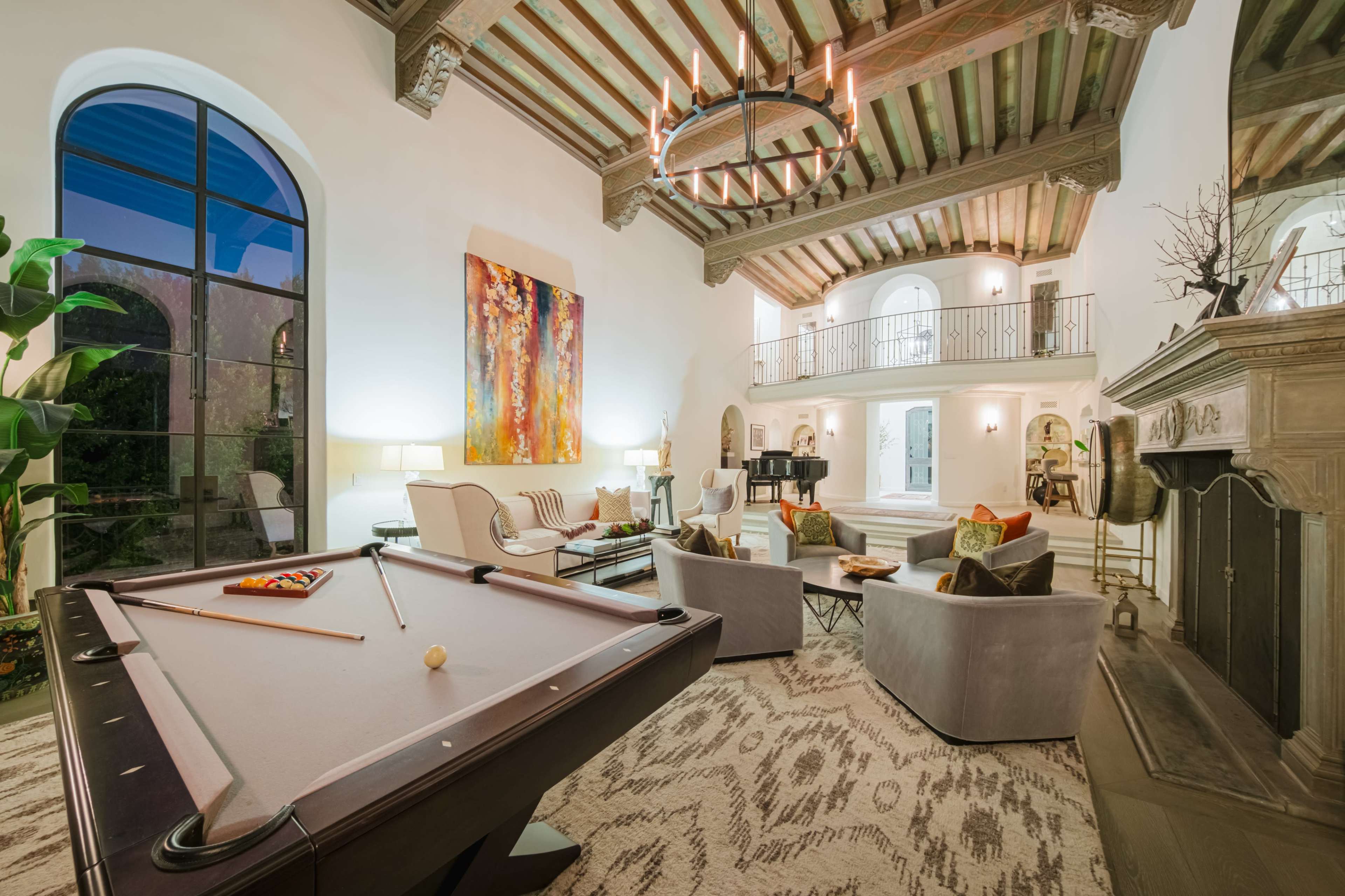 The image shows a spacious, elegantly furnished living area with a pool table in the foreground, high wooden ceilings, and large windows allowing natural light to flow in.