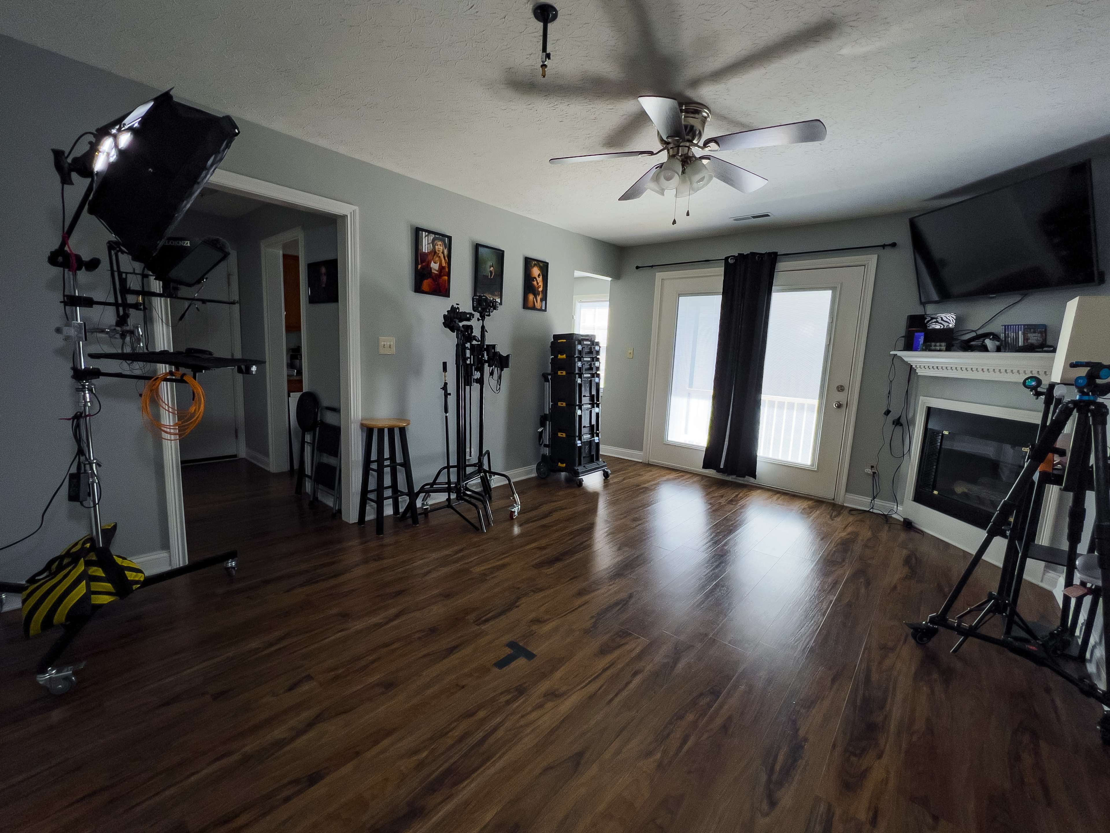 The image shows a spacious room with a wooden floor, featuring photography equipment on stands, a fireplace, and large windows with curtains.