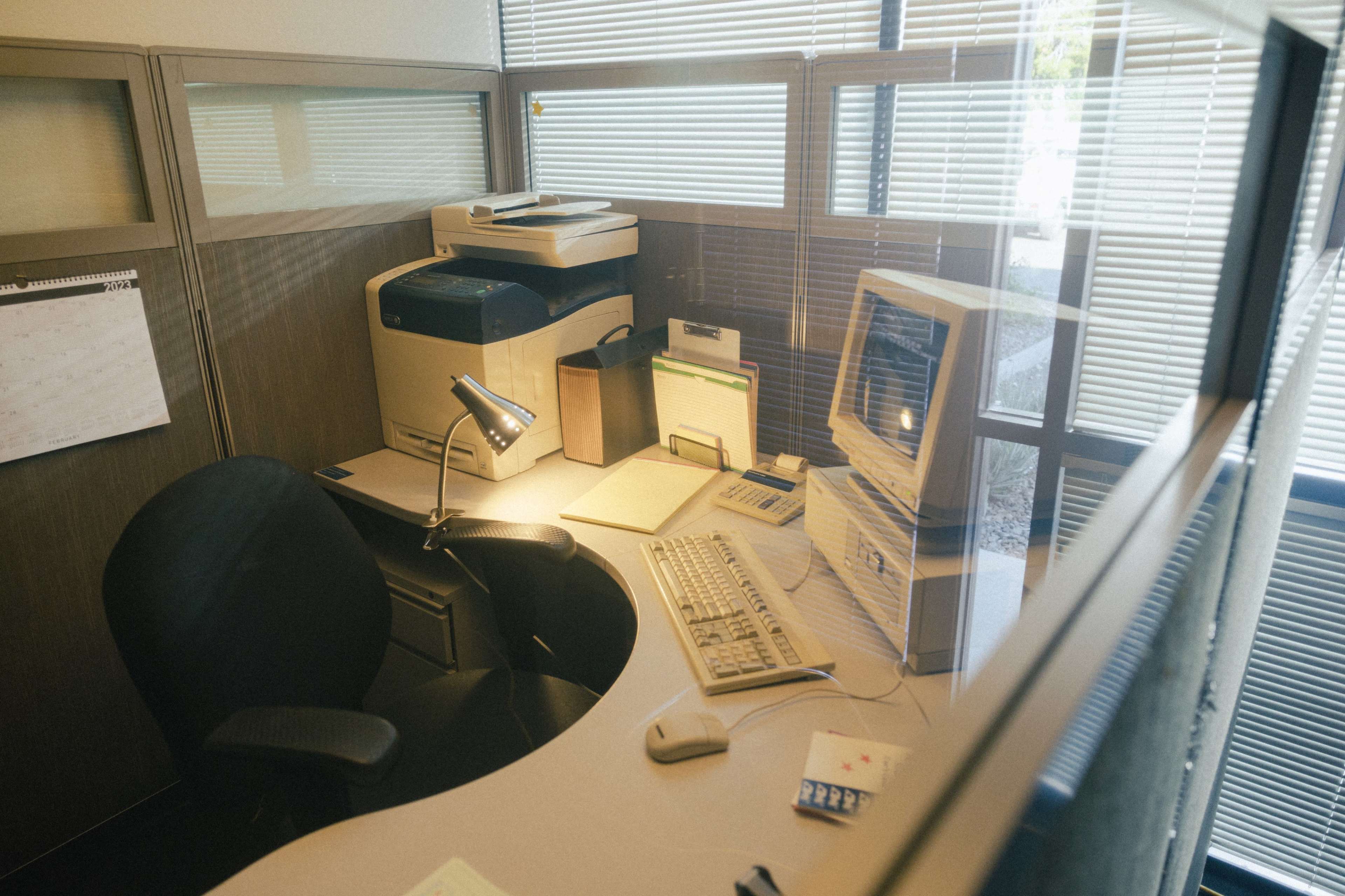 An office space features a desk with a computer, printer, lamp, and various office supplies, enclosed by glass partitions.