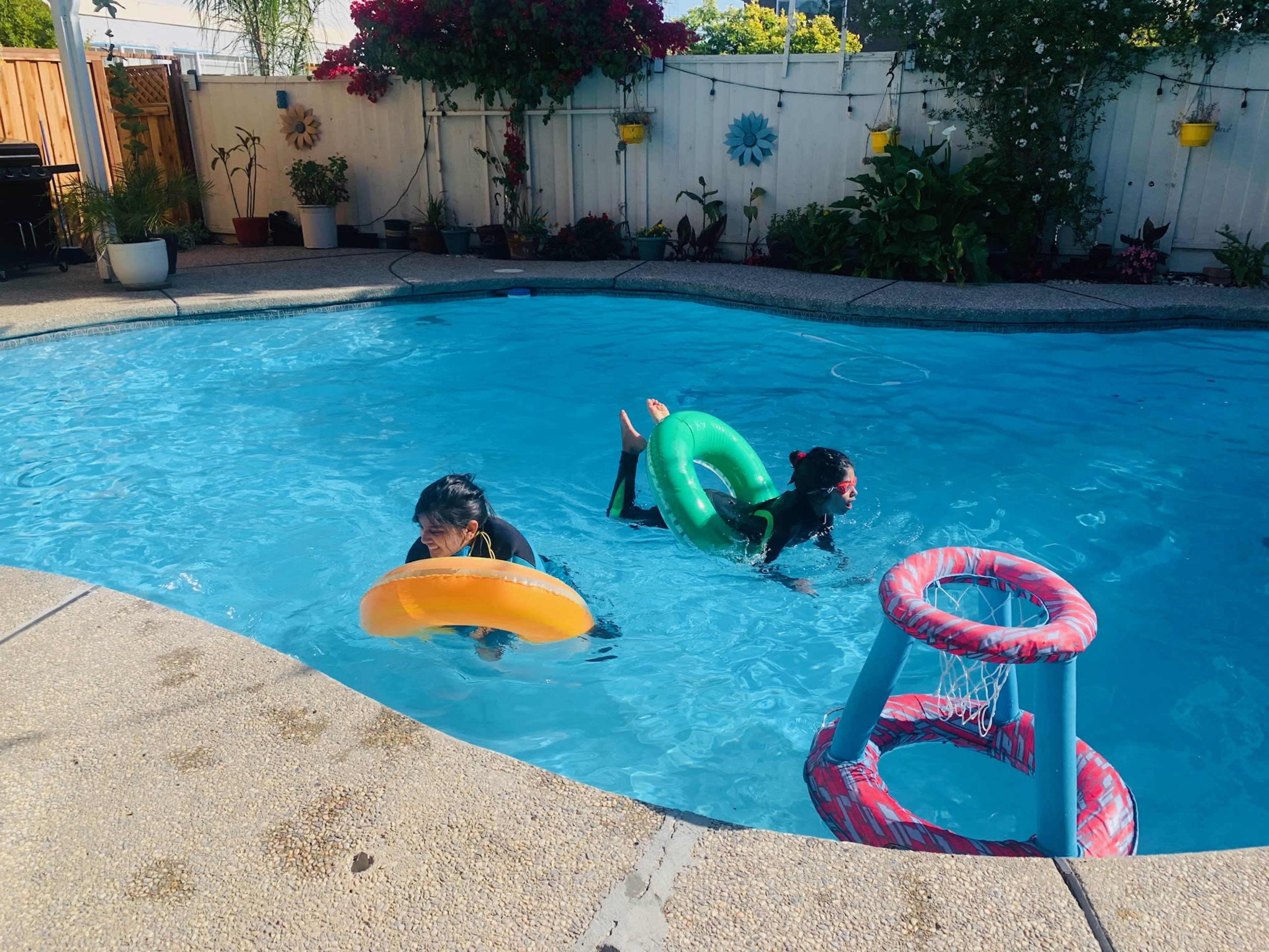 Two children play in a swimming pool, one holding an orange float and the other using a green float, while a basketball hoop stands at the pool's edge.