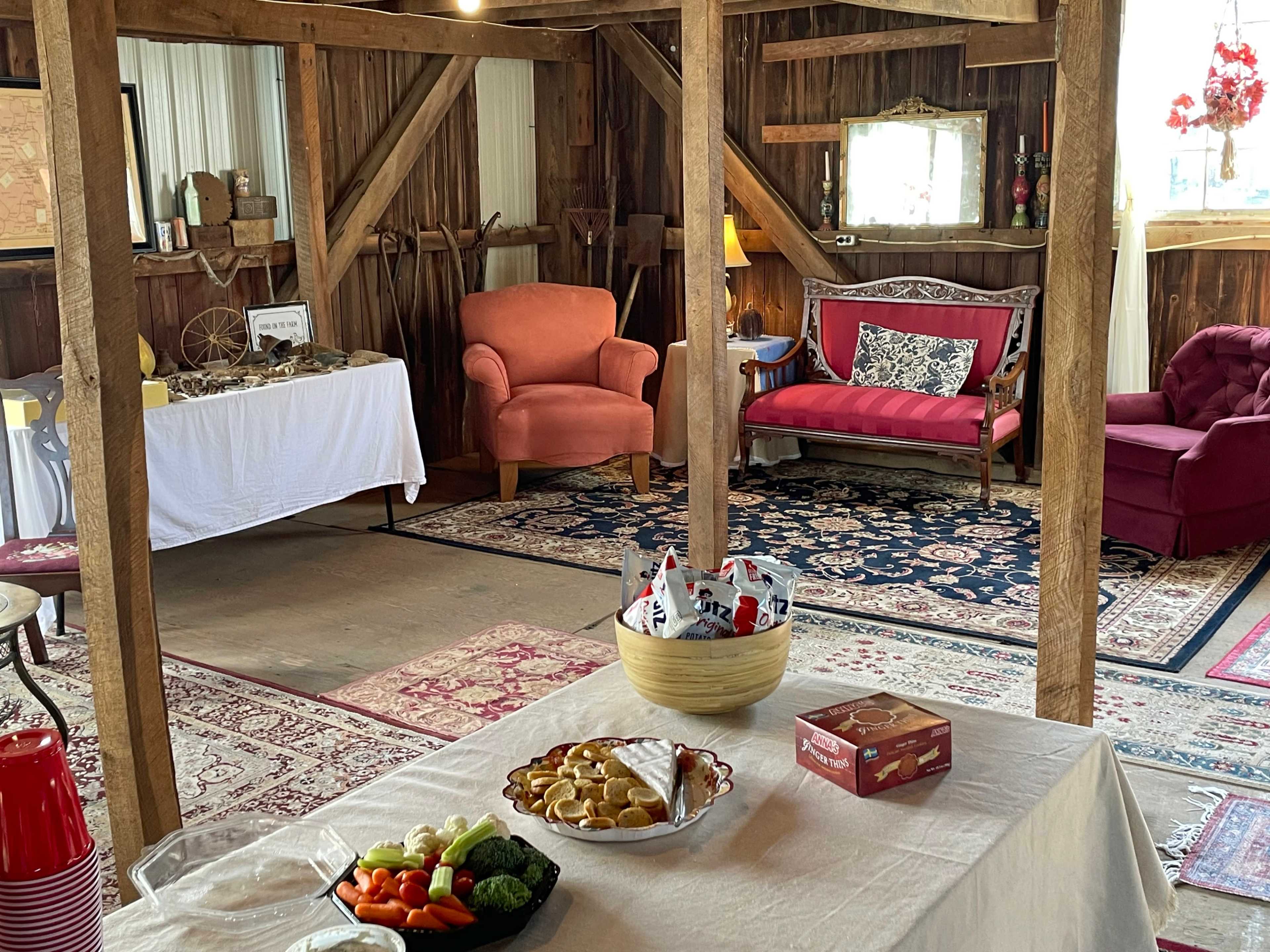 A rustic interior space with wooden beams, two sofas, a table set with a platter of vegetables and snacks, and a white tablecloth.