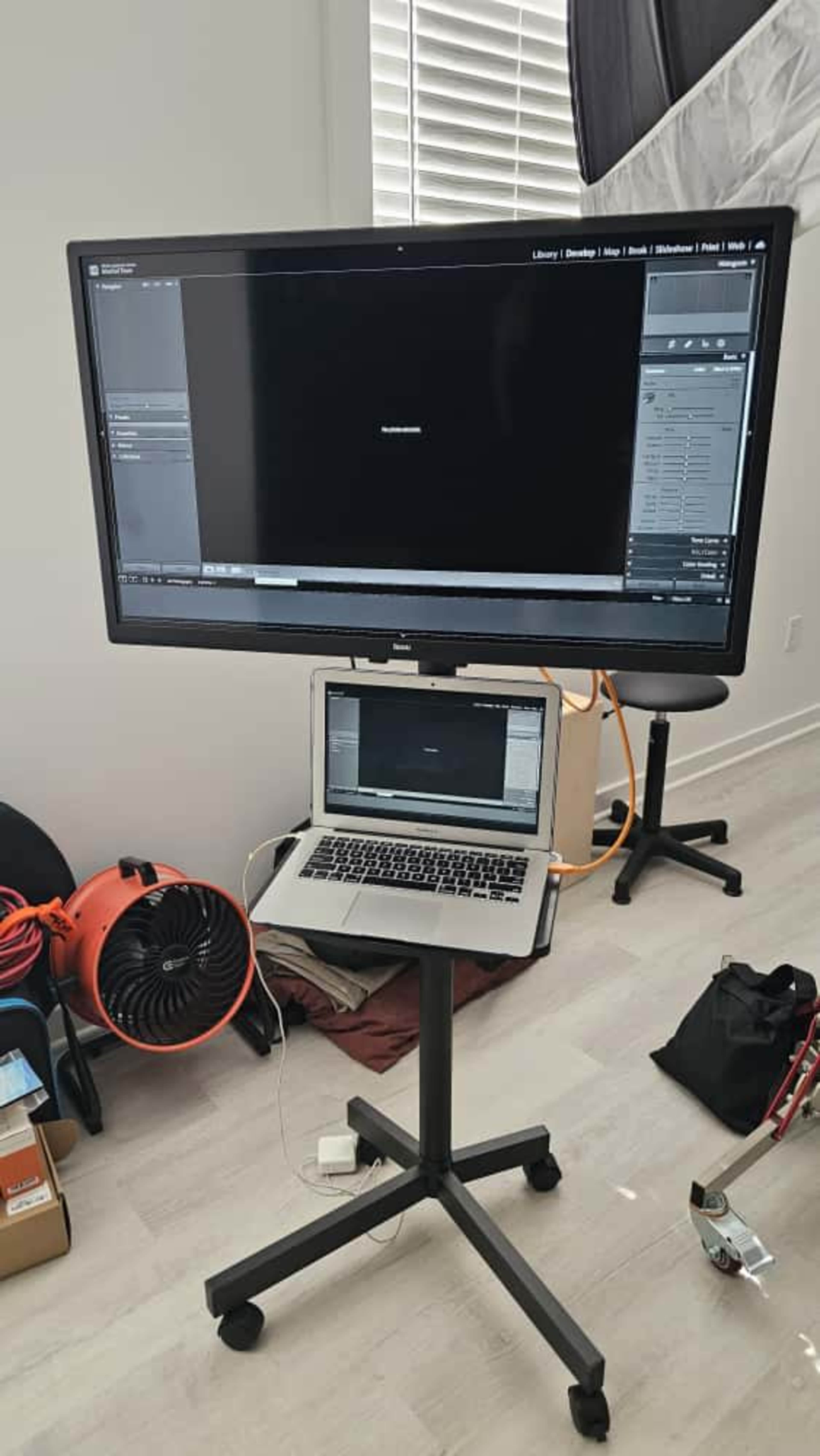 A large monitor is mounted on a mobile stand next to a laptop, with various equipment and tools visible in the background.