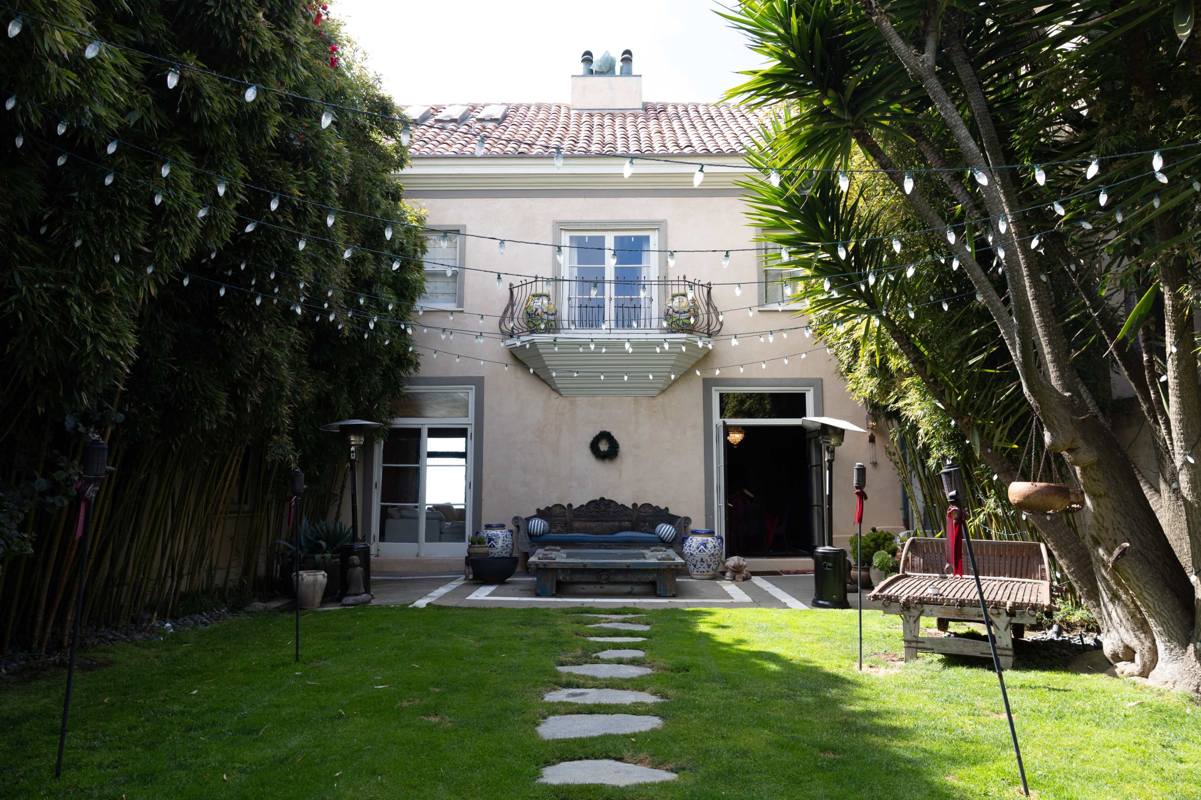 A well-maintained backyard features a stone path leading to a house with a balcony, surrounded by greenery and string lights.