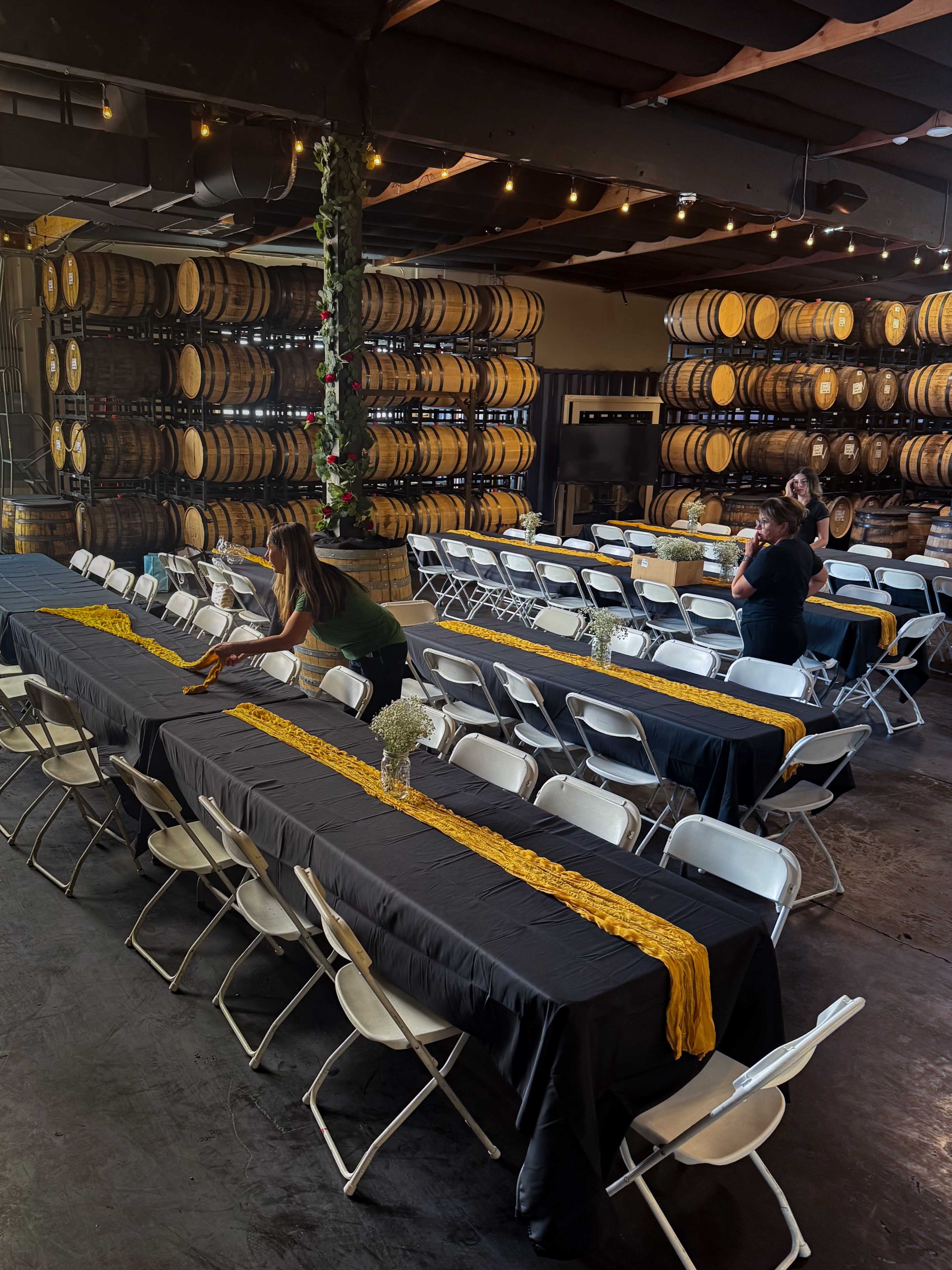 A spacious venue features long tables set with black tablecloths and yellow runners, surrounded by barrels stacked on shelves in the background.