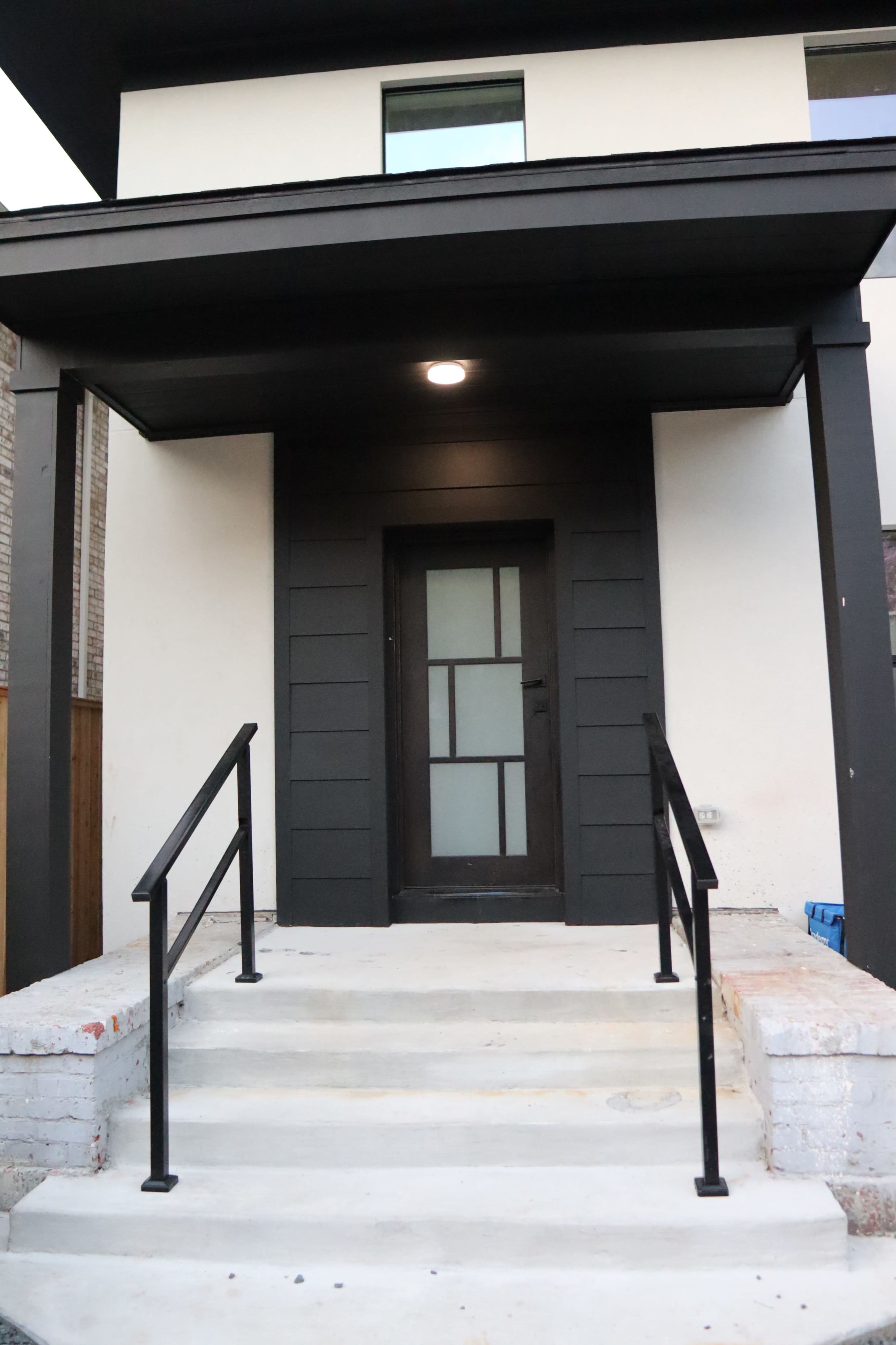 The entrance of a modern house with a dark front door and metal railings leading up to a set of concrete steps.