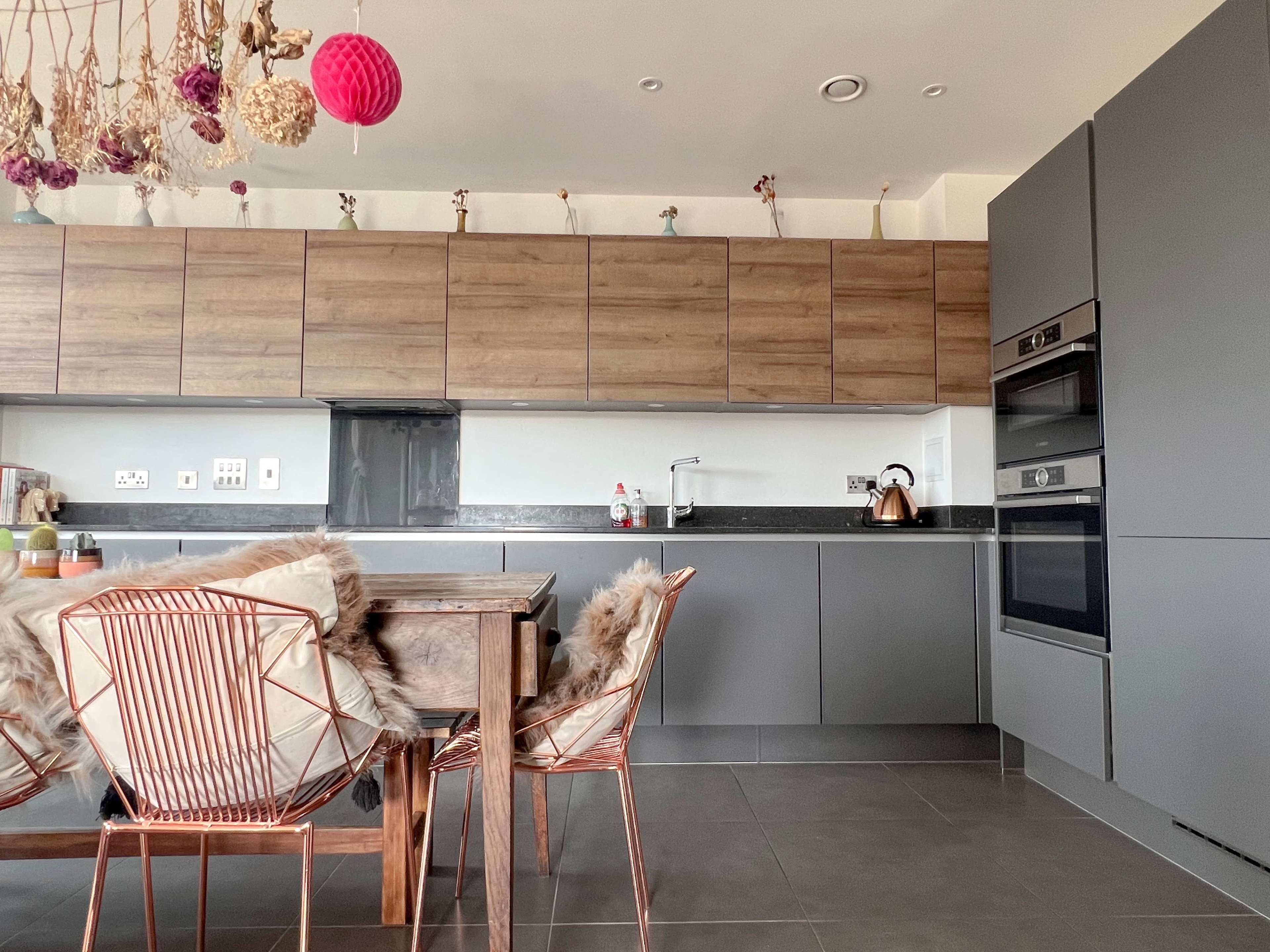 A modern kitchen featuring wooden cabinets, grey appliances, a dining table with copper-colored chairs, and decorative elements hanging from the ceiling.