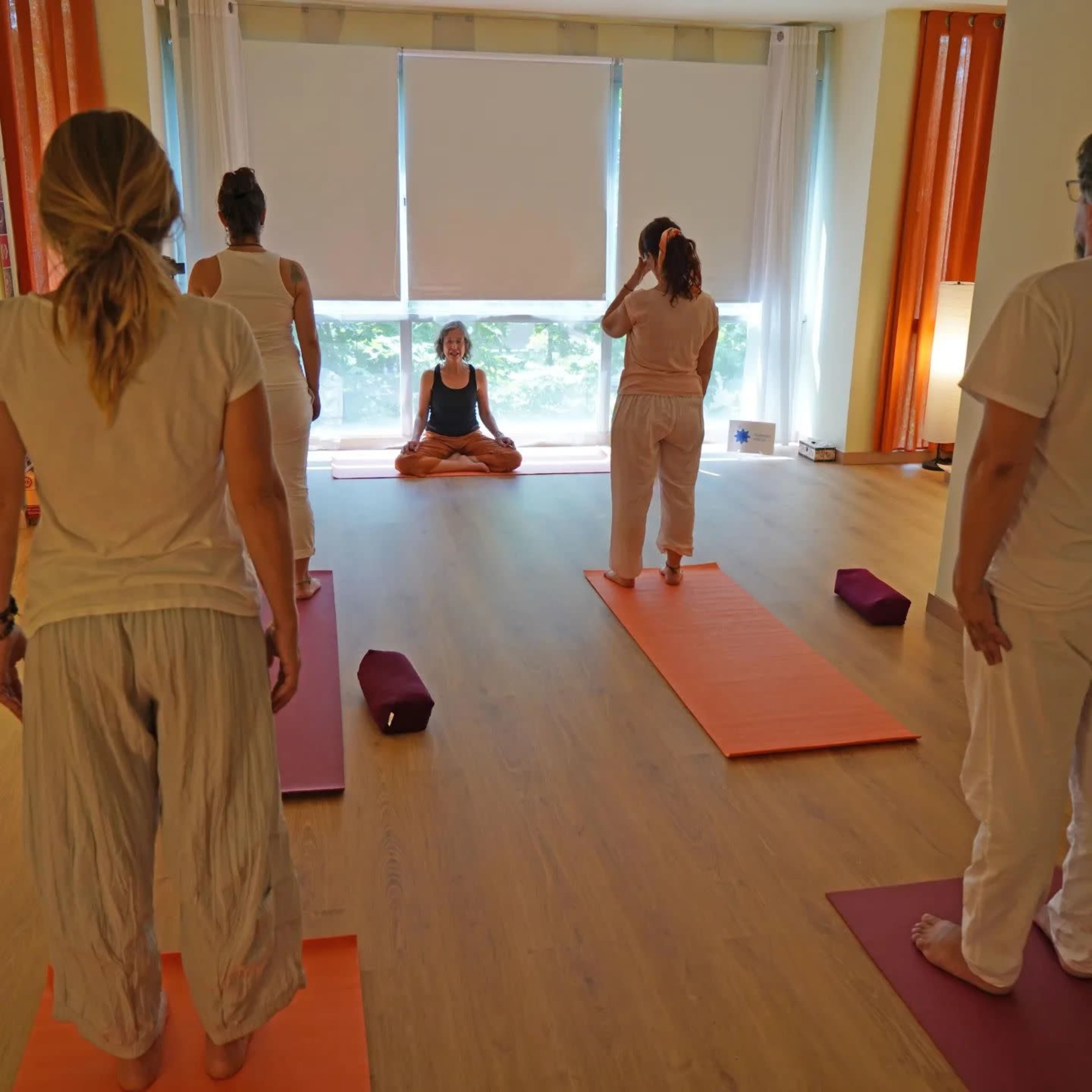 A group of people practices yoga on orange mats in a brightly lit room, facing an instructor seated at the front.
