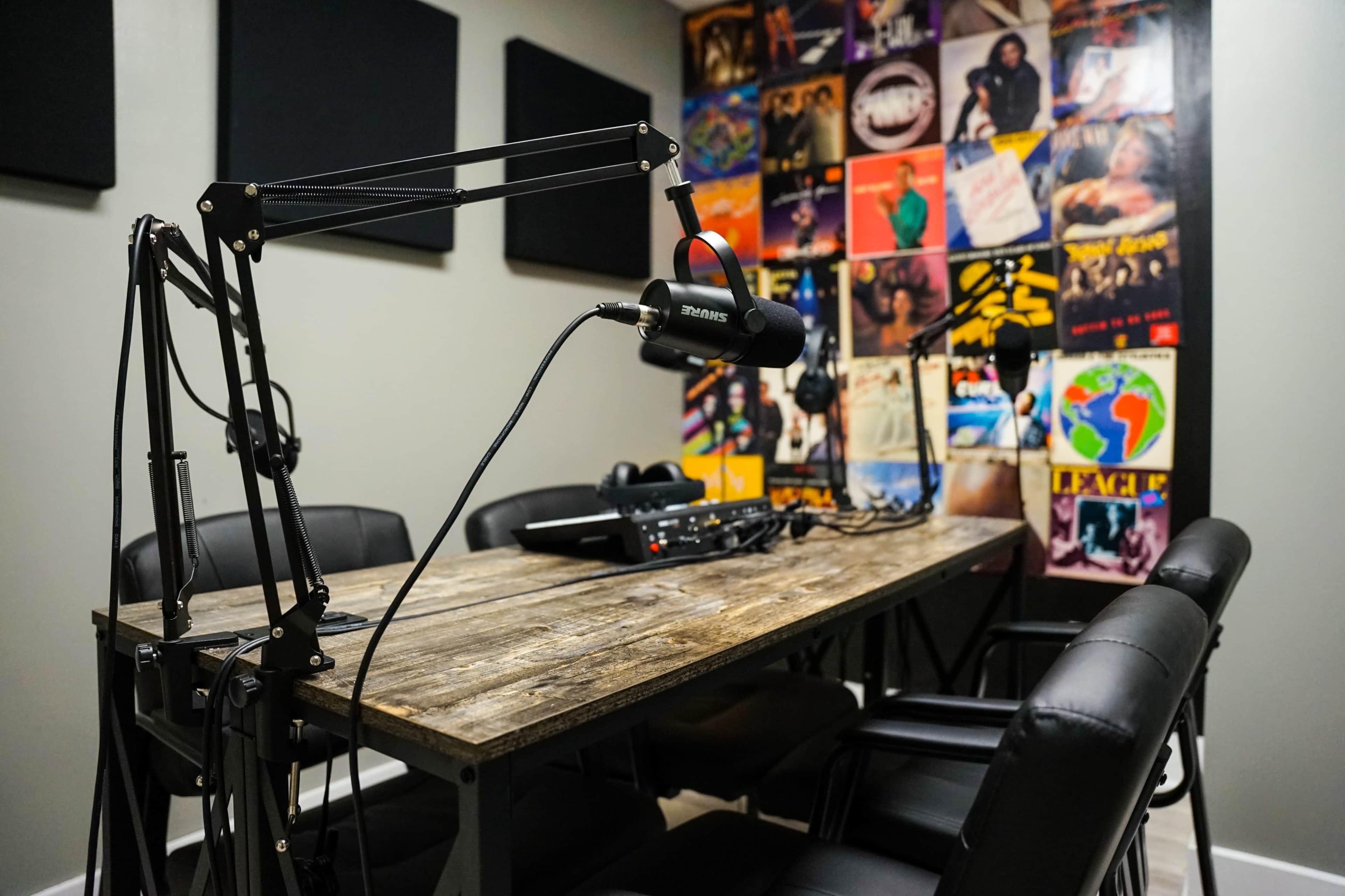 A recording studio features a wooden table with microphones, a mixer, and black chairs, set against a wall adorned with colorful vintage vinyl records.