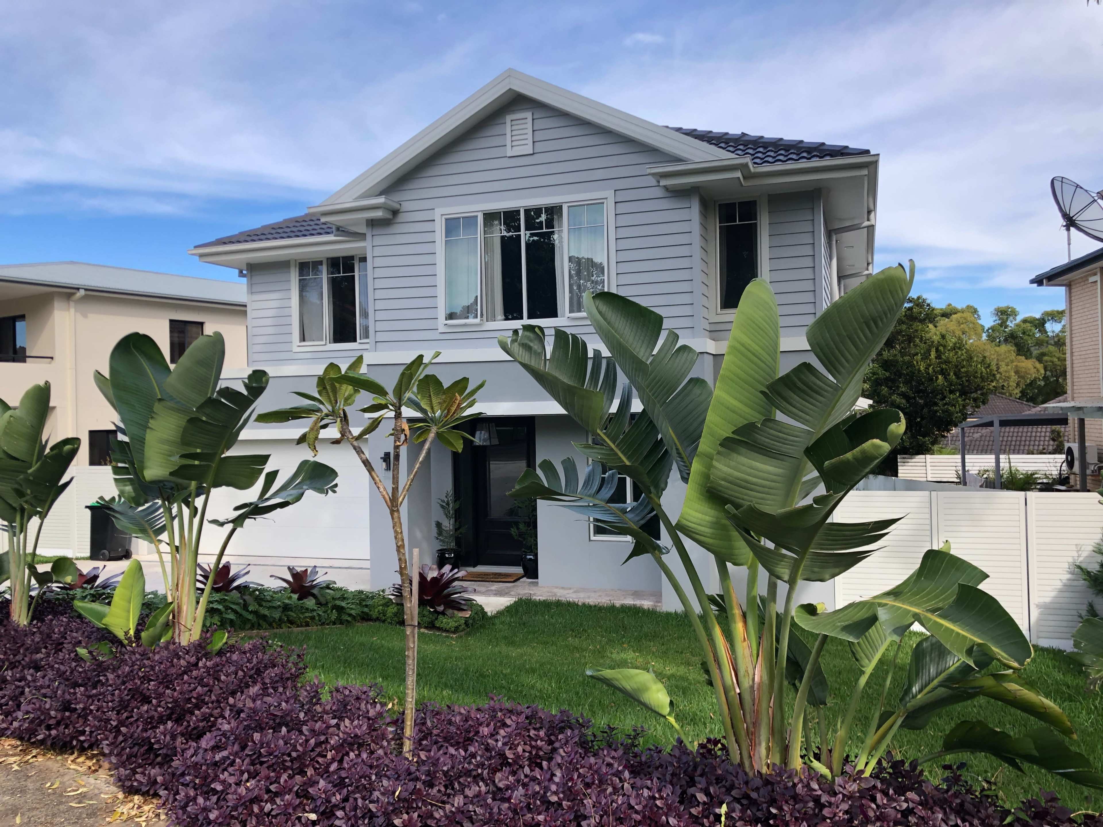 A two-story gray house with large windows is surrounded by tropical plants and a well-maintained garden.
