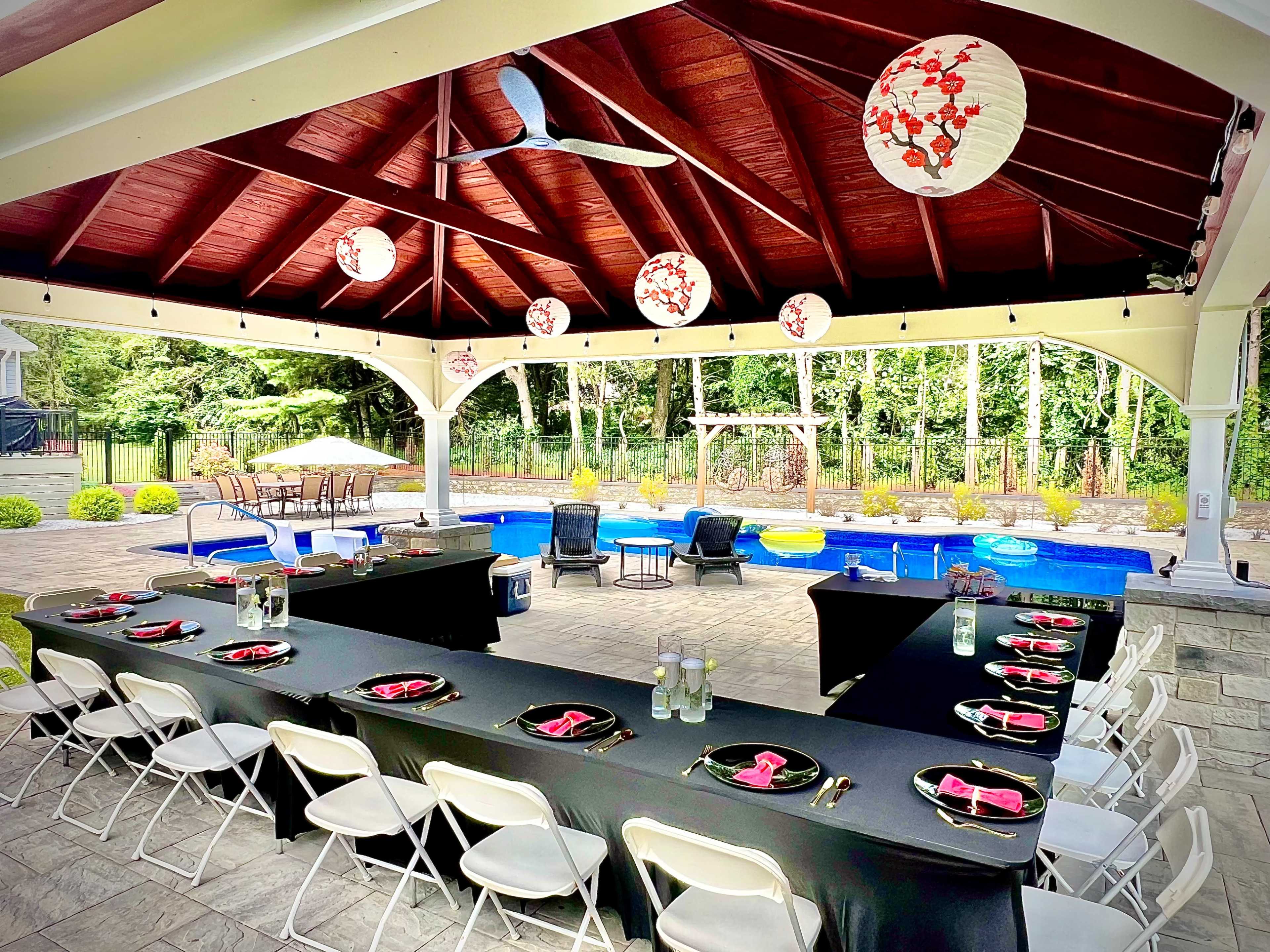 A large outdoor pavilion with a dining table set up beside a pool, decorated with lanterns and surrounded by lush greenery.