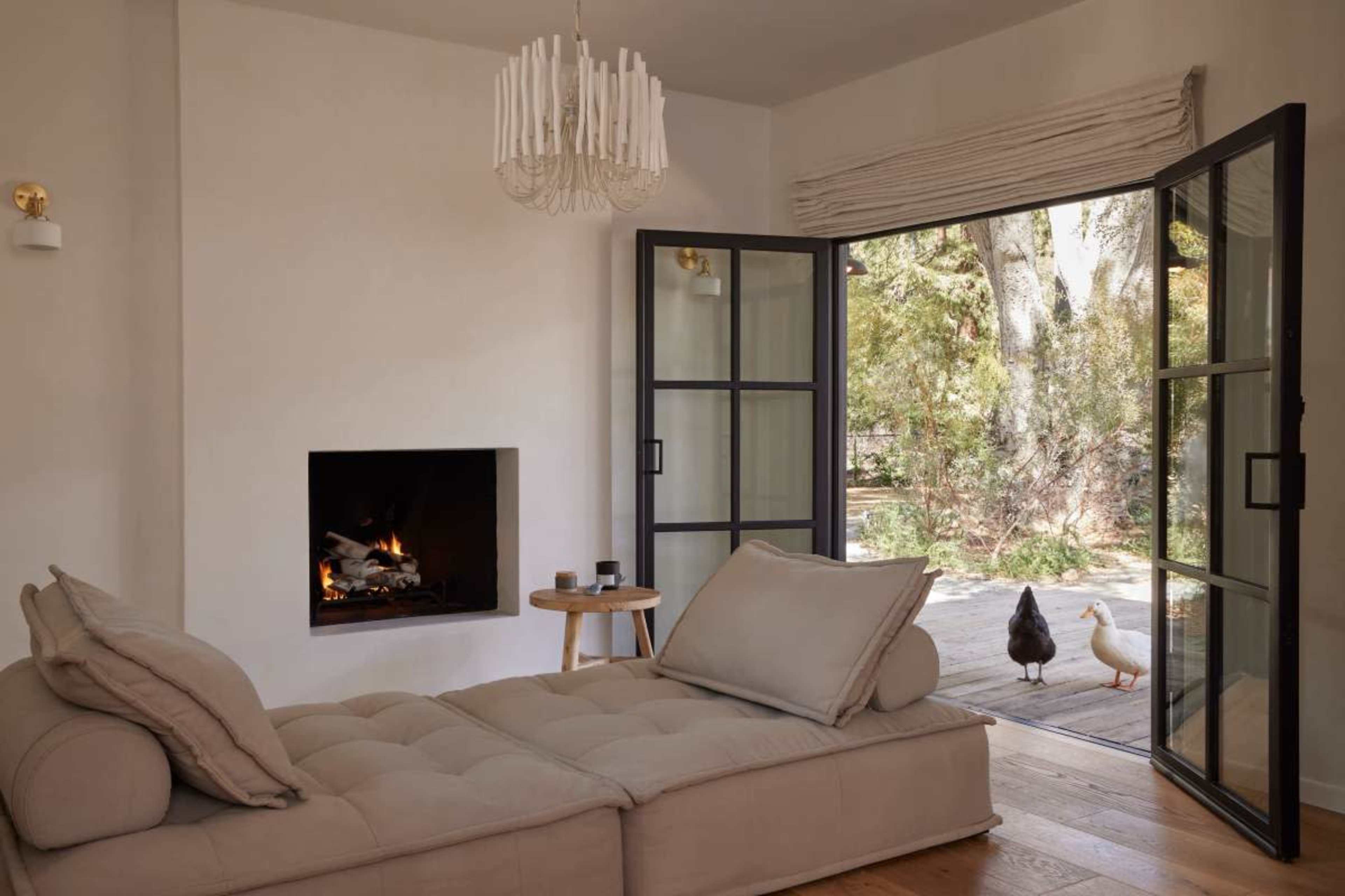 A cozy living room features a chaise lounge facing a fireplace, with large open doors leading to an outdoor area where two ducks are visible.