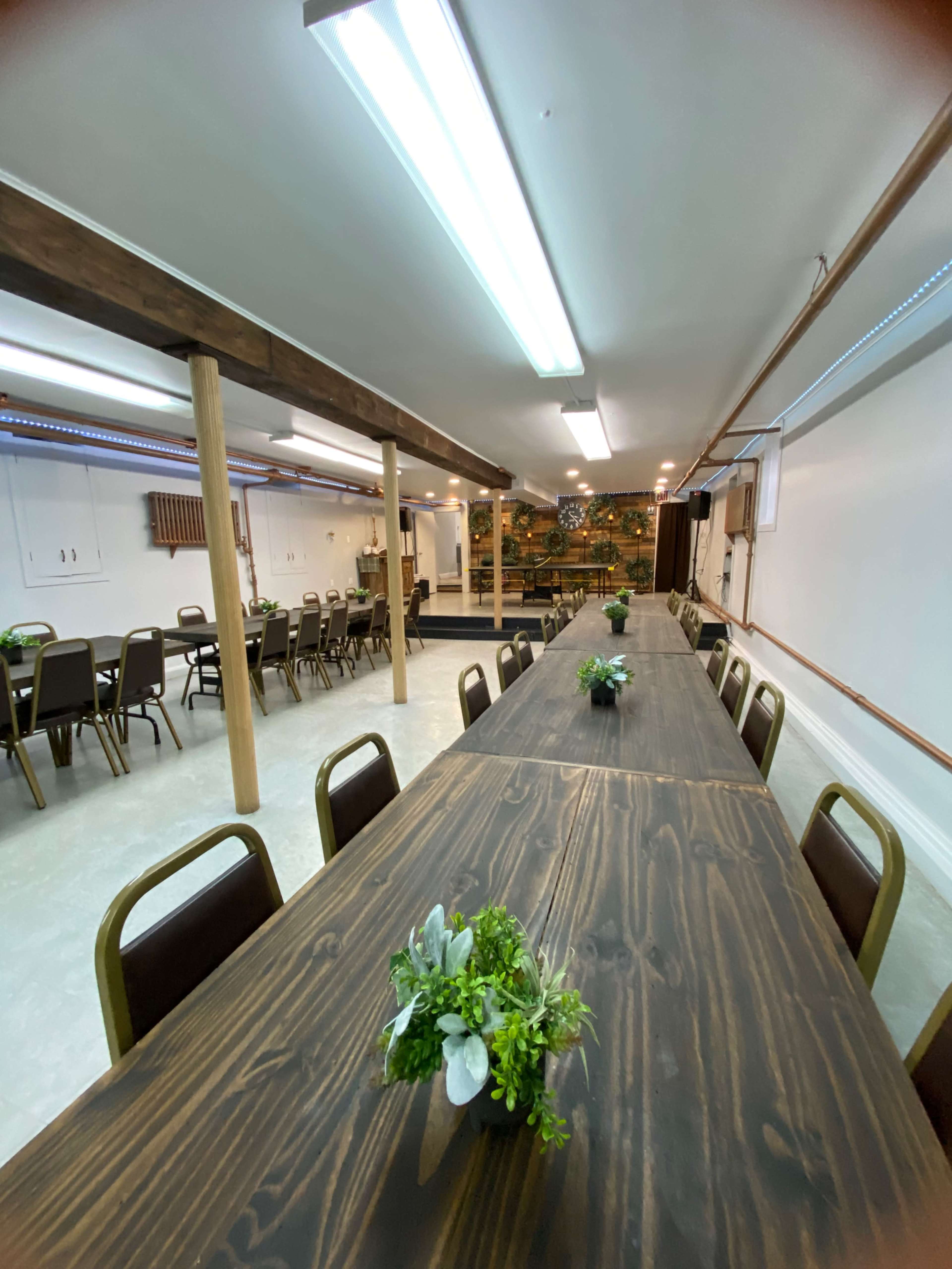 A spacious indoor room with two long wooden tables set up for seating, each adorned with small potted plants, and additional seating arranged along the sides.