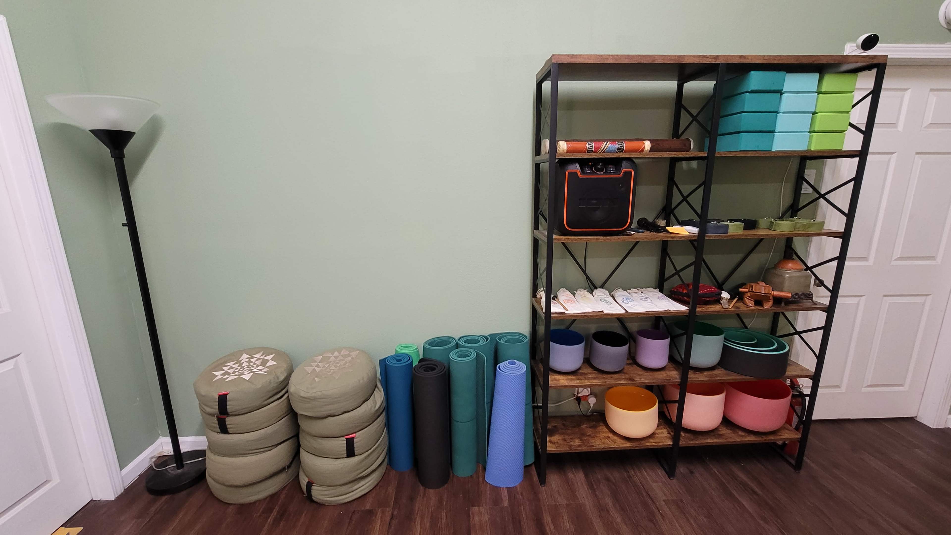 A shelf filled with various containers and yoga supplies stands against a green wall, next to stacked meditation cushions and yoga mats.