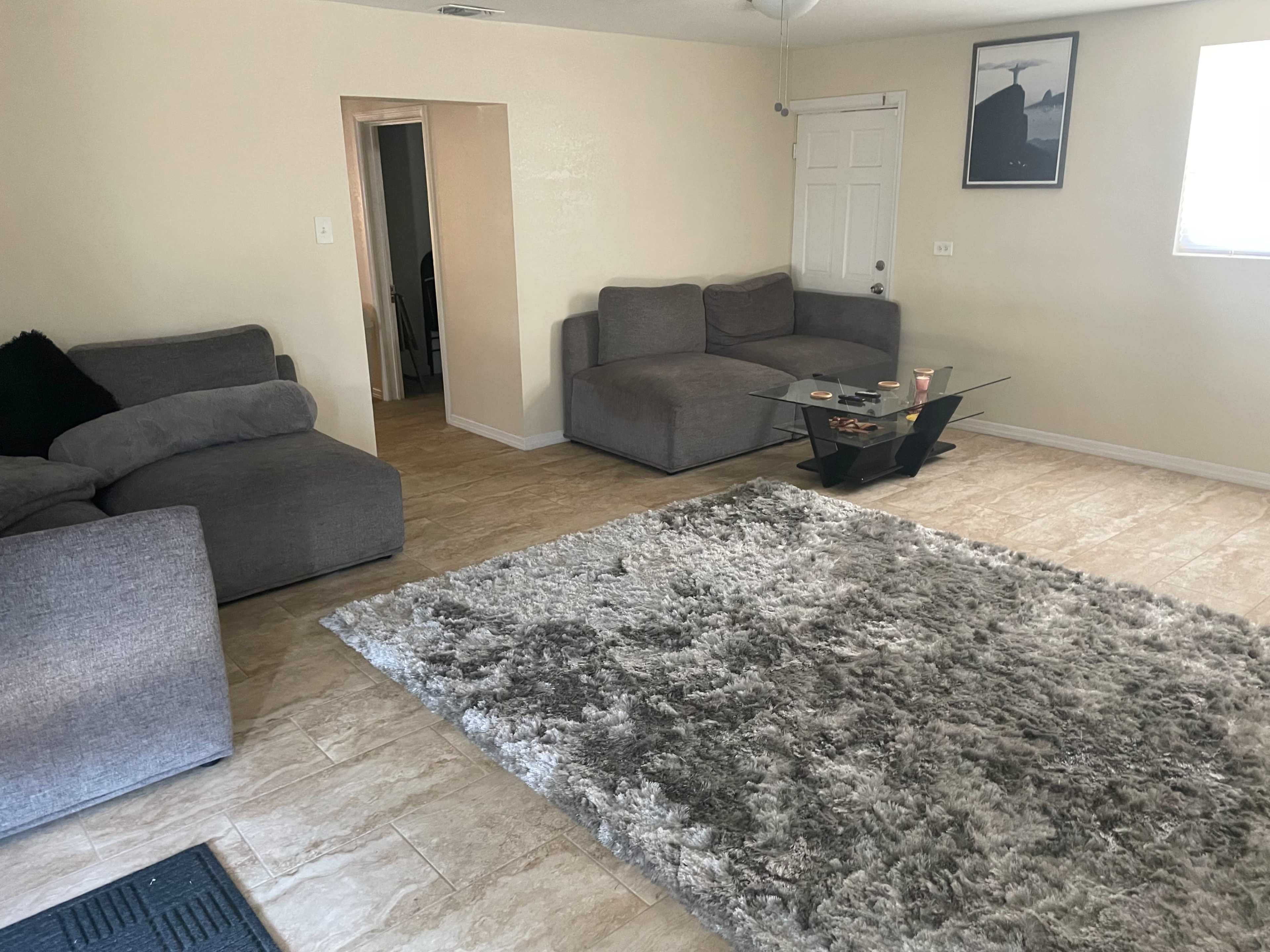 A living room with gray sofas, a glass coffee table, and a large area rug on a tiled floor.