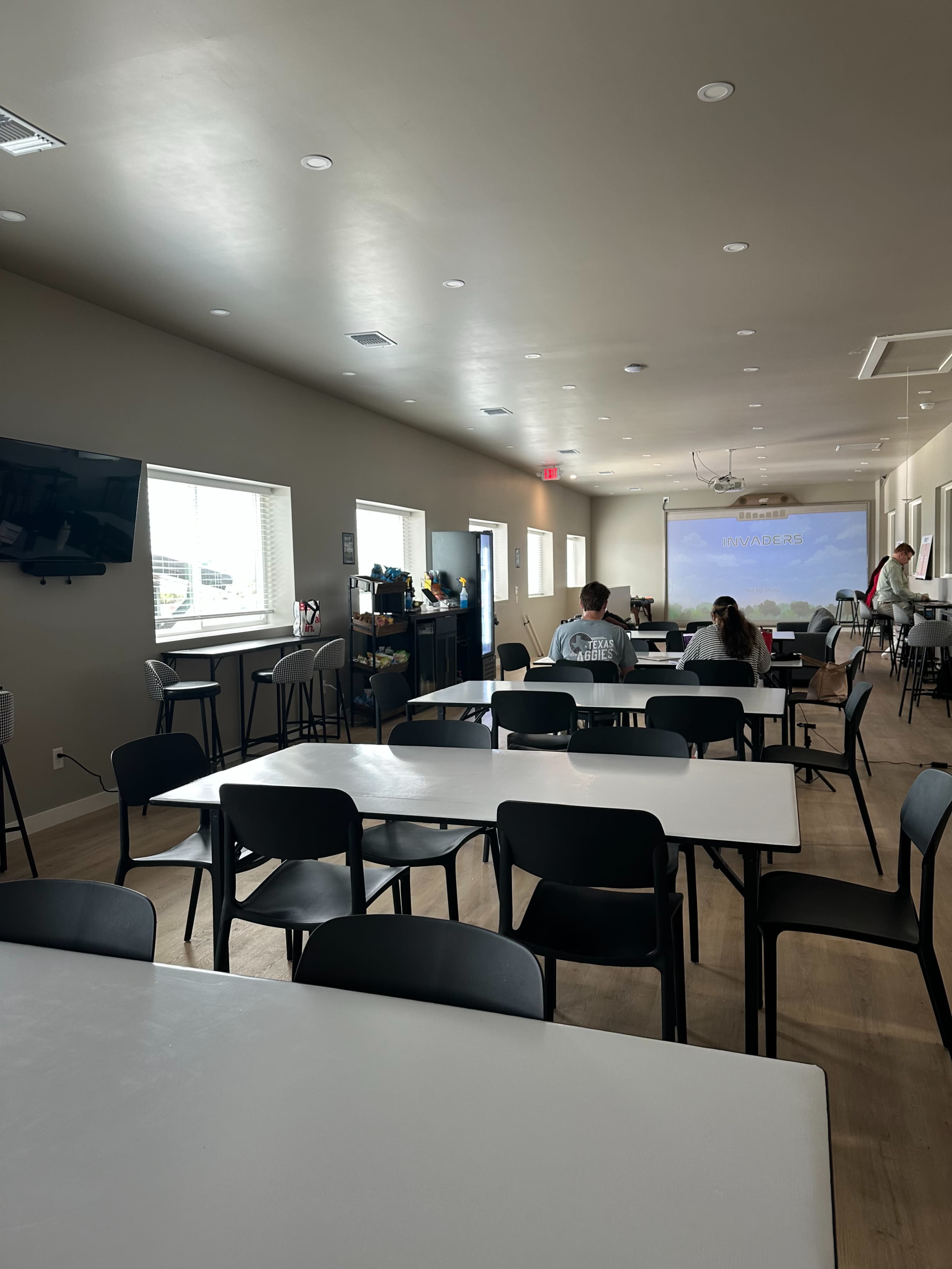 The image shows a spacious, empty room with a few tables and chairs, and a projector screen displaying a presentation at the far end.