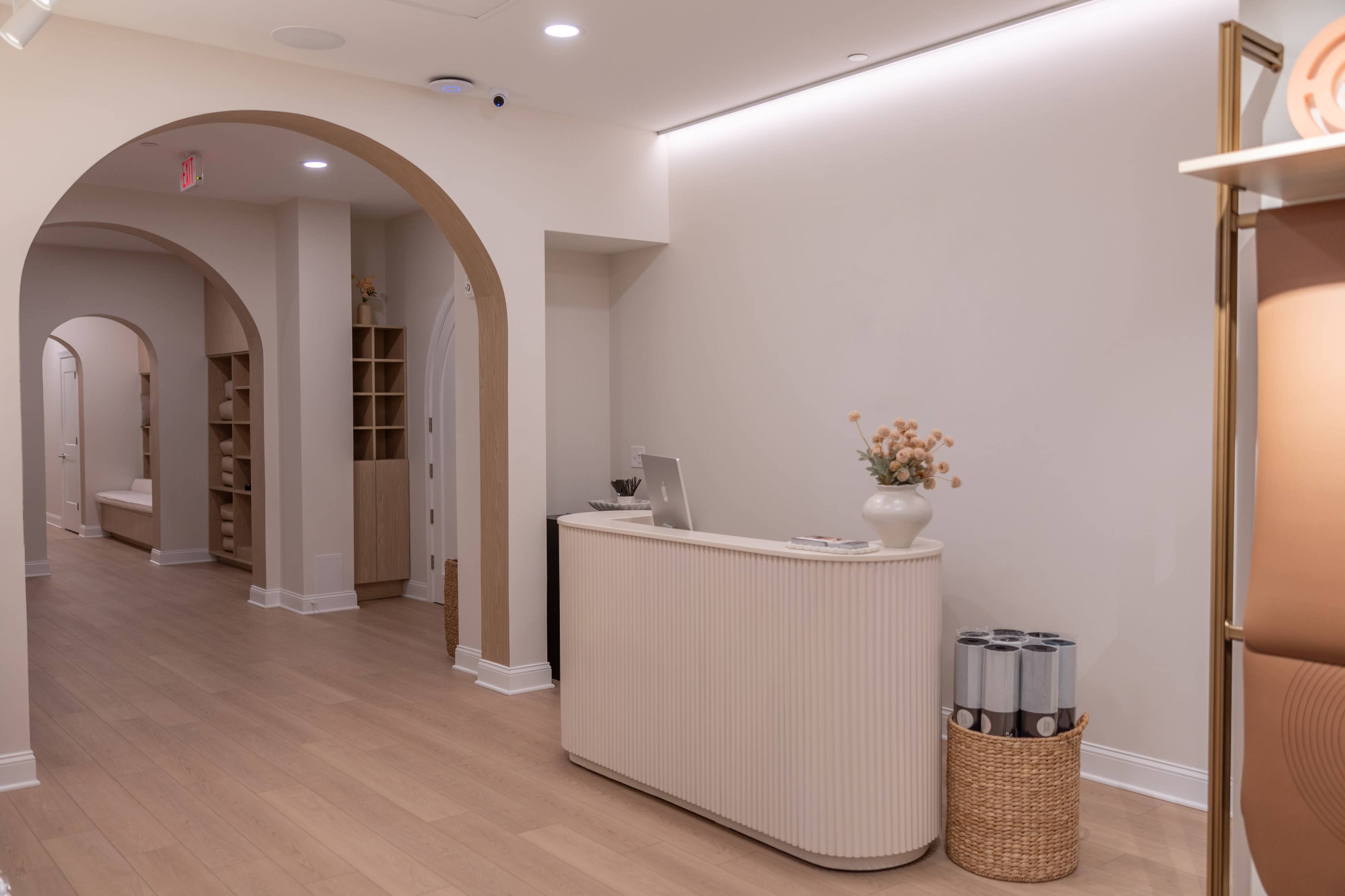 A reception area features a curved front desk with decorative items, surrounded by soft arches and light wood flooring.