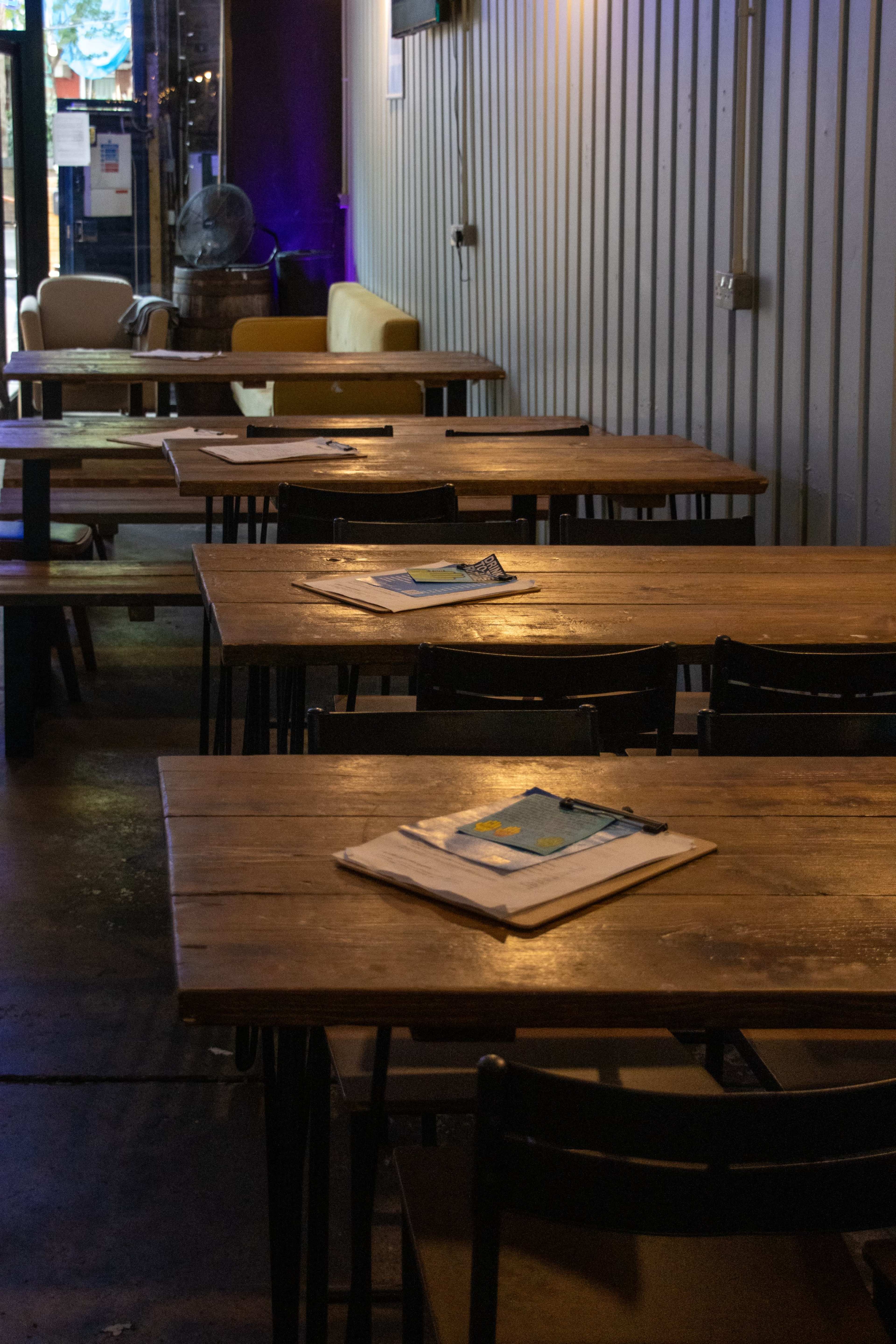 The image shows a row of wooden tables in a dimly lit space, each with a stack of paper and a pen placed on top.