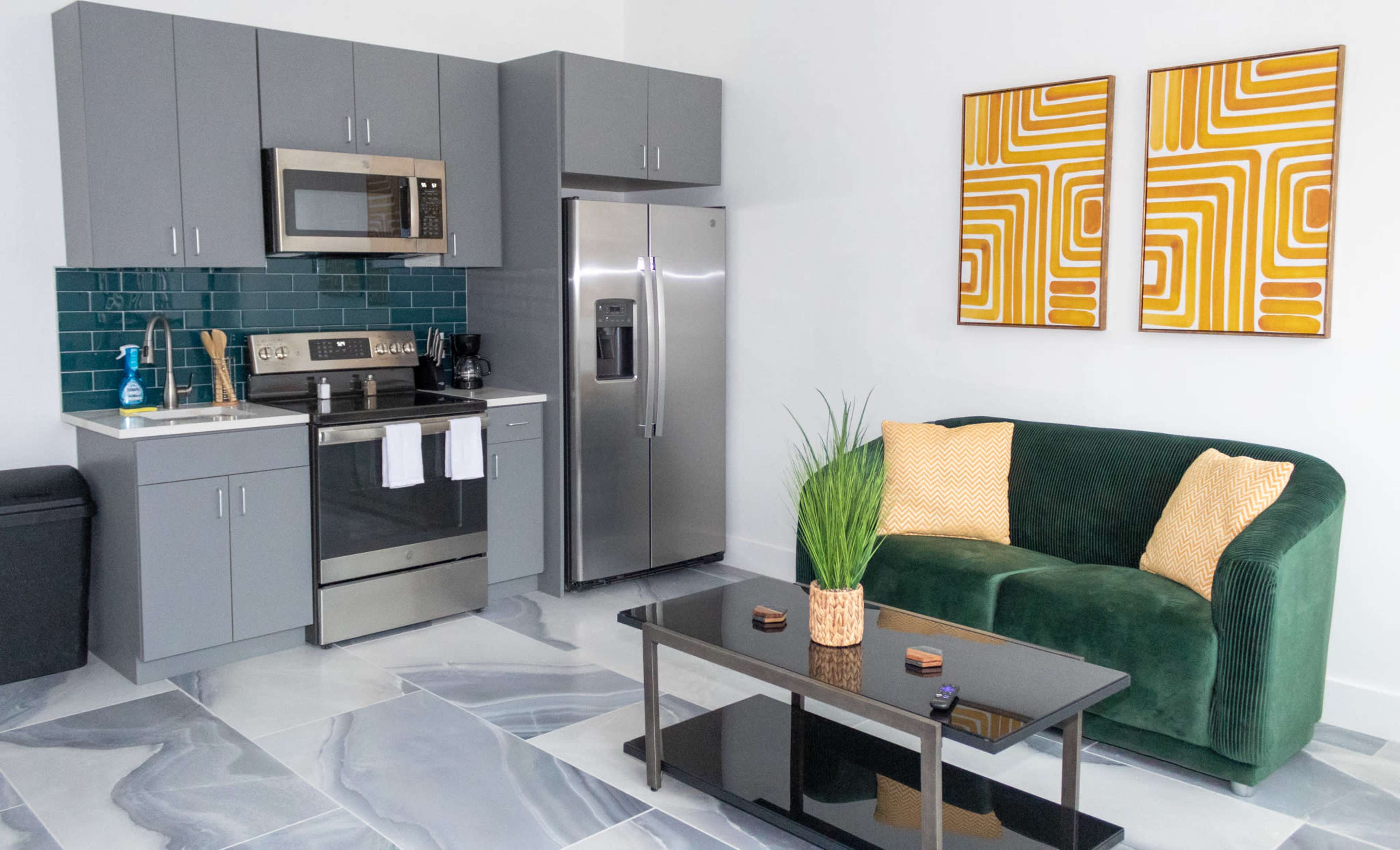 A modern kitchen and living area that features gray cabinets, stainless steel appliances, a green sofa, and abstract wall art.