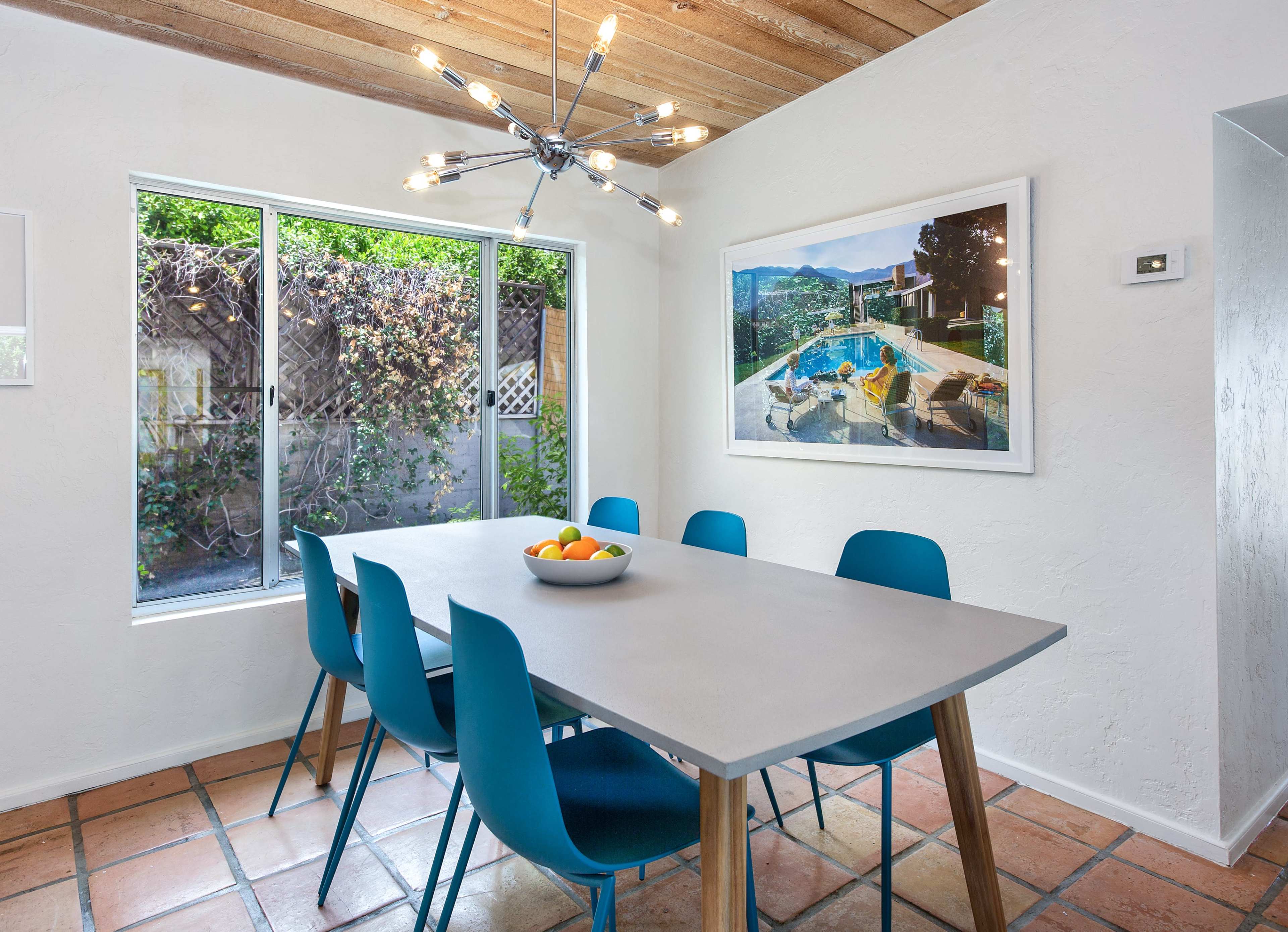 A dining area features a long table surrounded by blue chairs, a large window overlooking a garden, and a modern light fixture above.