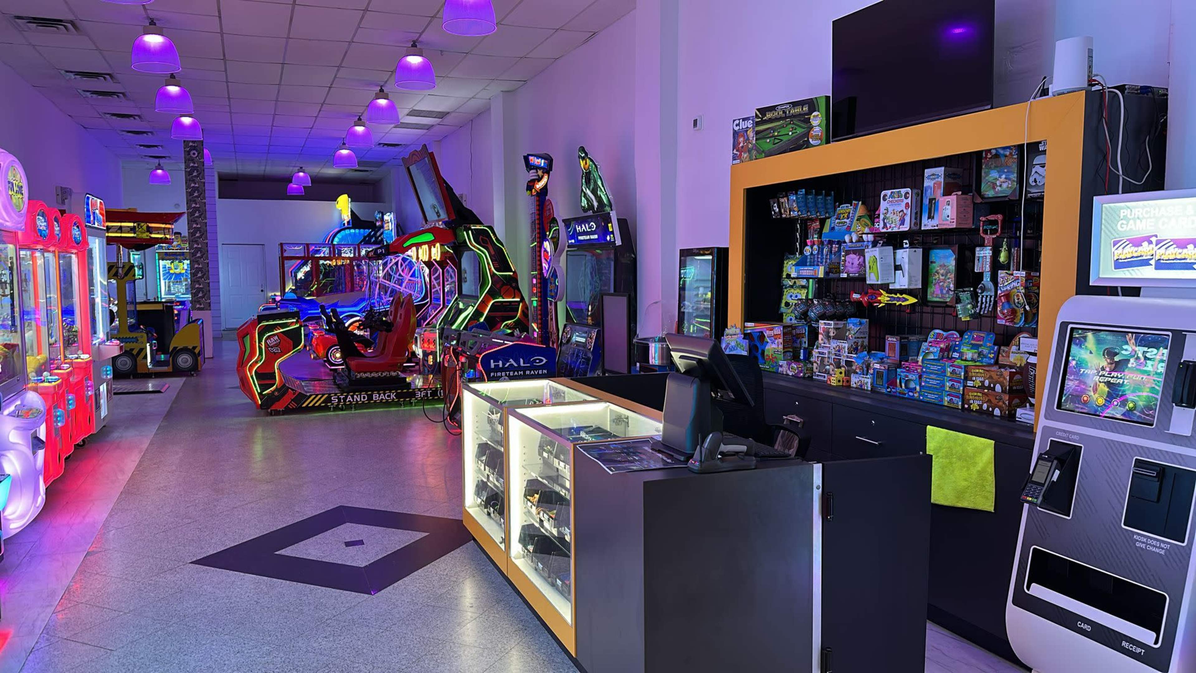 The image shows an arcade with various gaming machines on one side, a counter with merchandise and prizes, and vibrant lighting throughout the space.