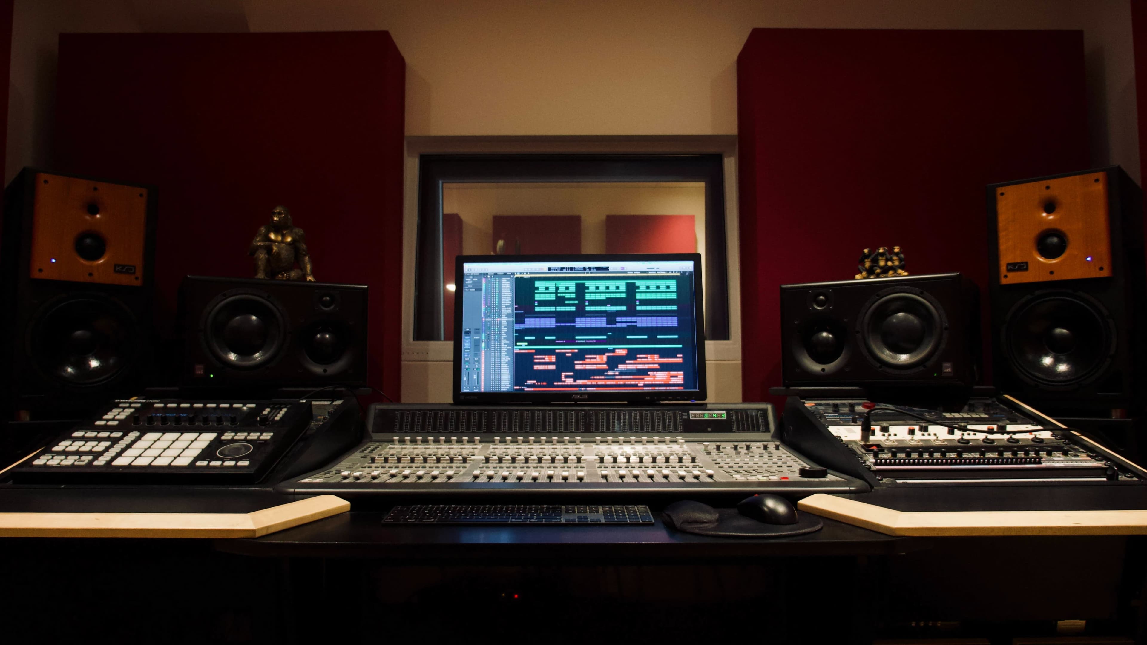 The image shows a music studio with a large mixing console, two computer screens displaying audio software, and various audio equipment on the desk and surrounding walls.