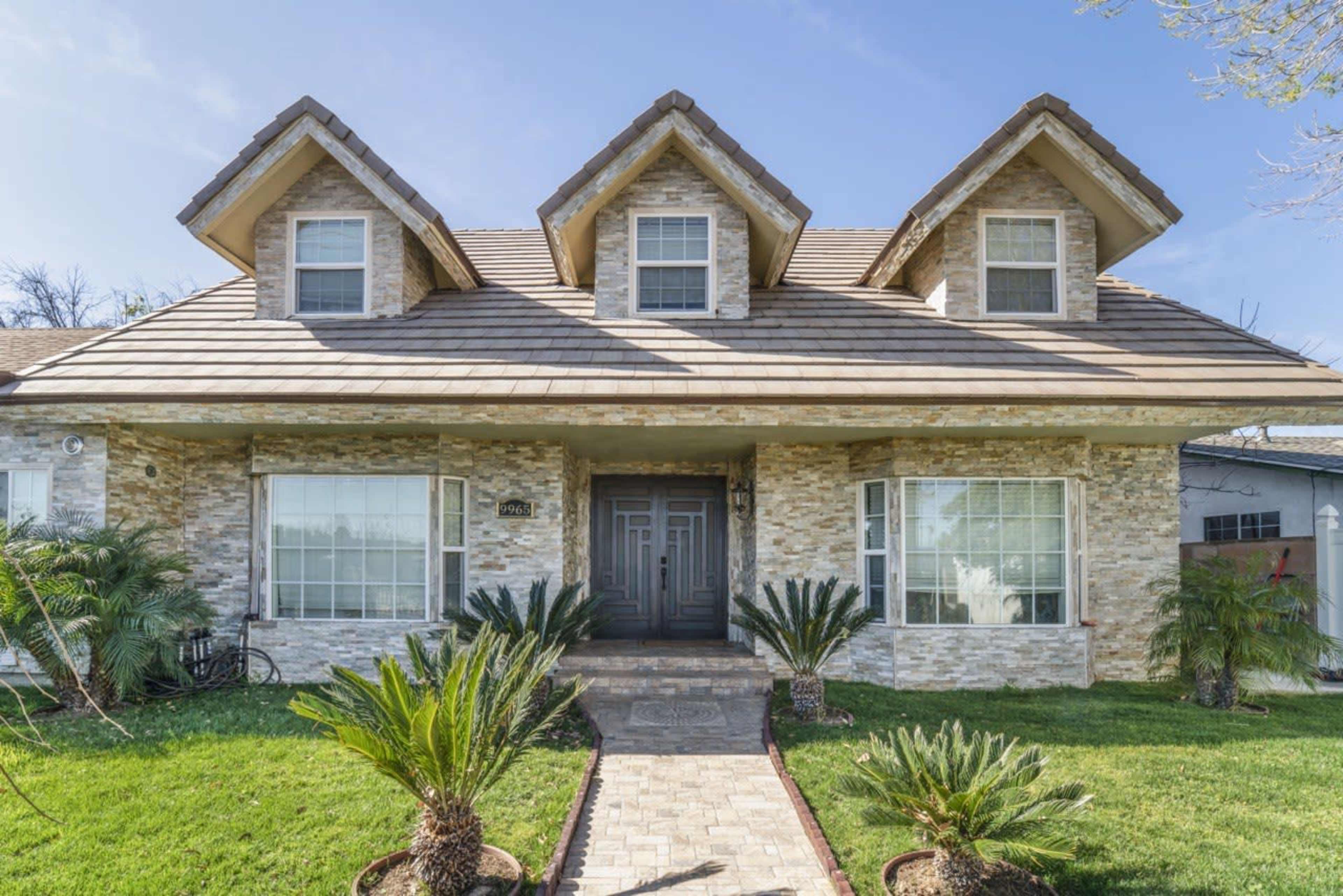 A two-story house with a stone facade and a gabled roof features a central entrance flanked by large windows and landscaped with palm plants.