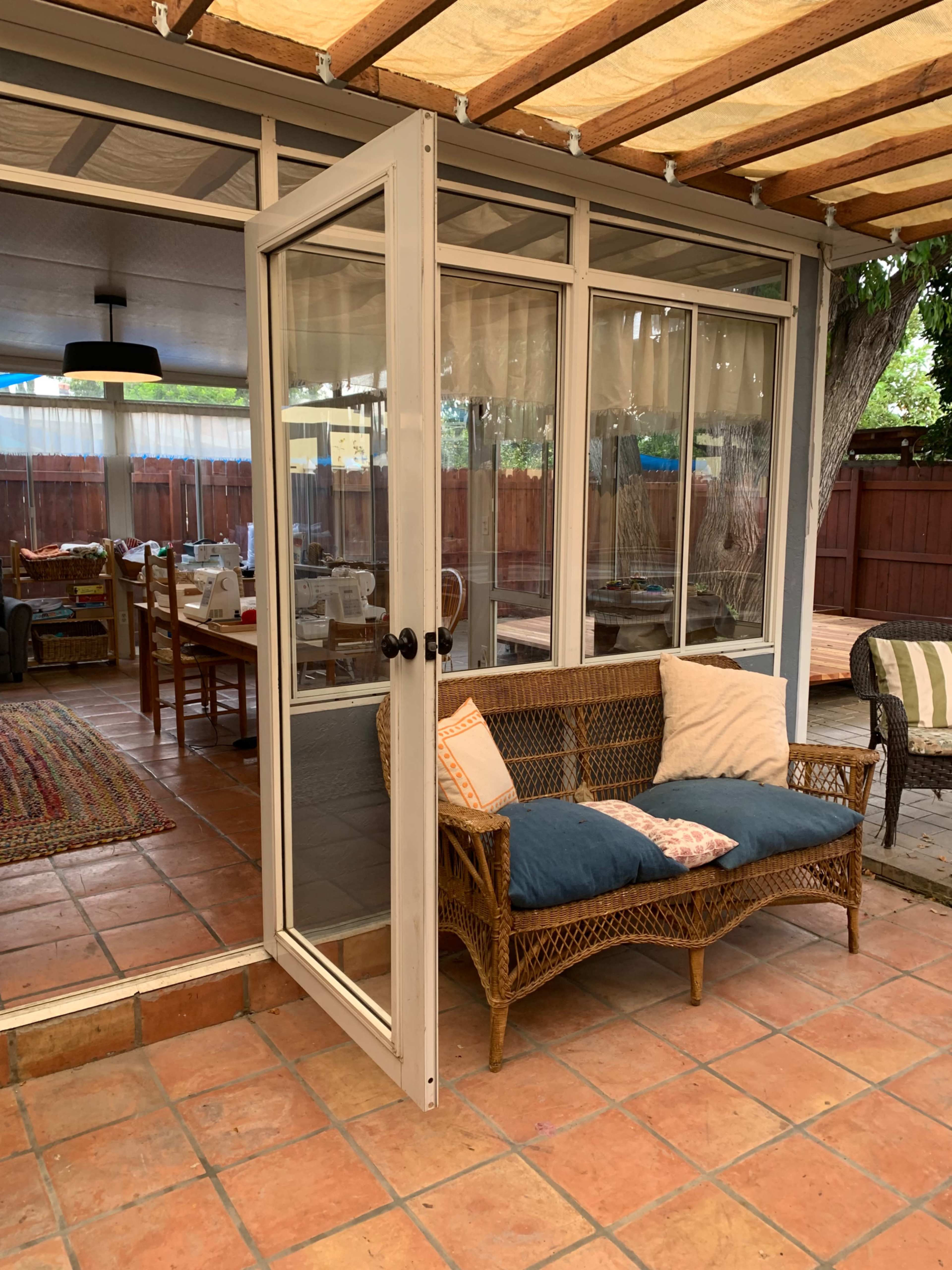 A cozy patio features a woven loveseat with blue cushions, surrounded by tiled flooring and glass doors leading to a dining area.