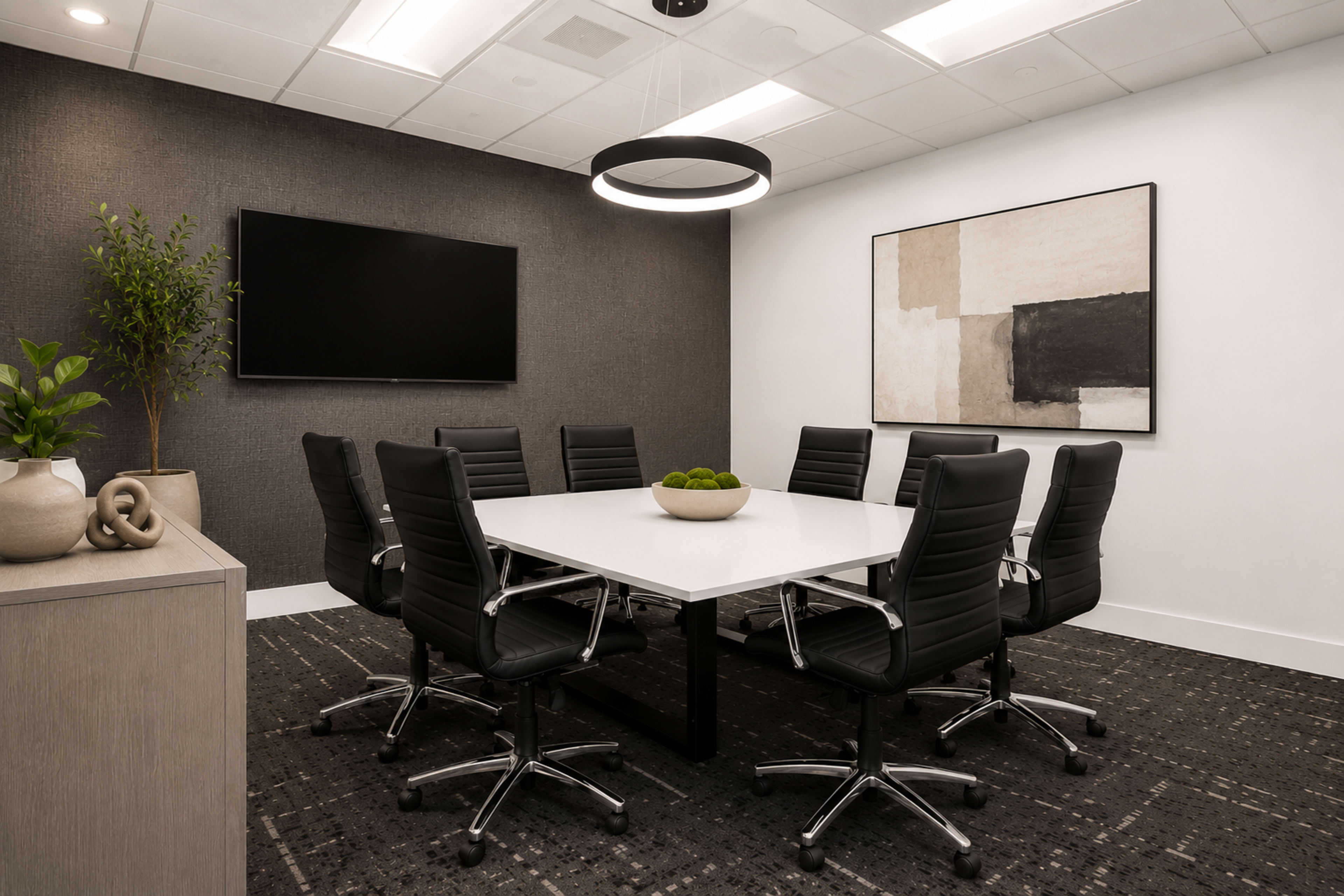 A modern conference room features a large white table surrounded by black chairs, with a television mounted on the wall and an abstract painting displayed nearby.