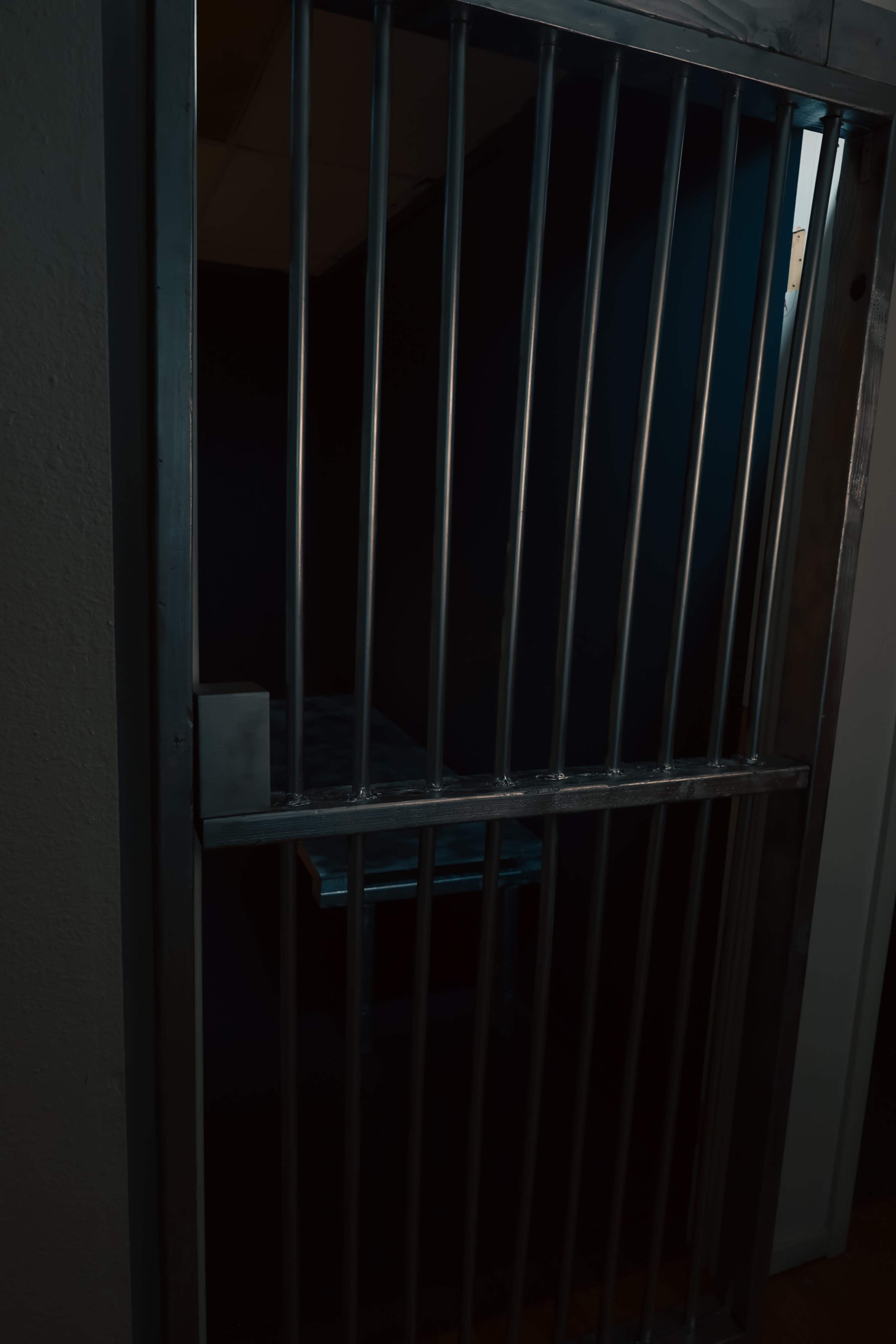 The image shows a partially open jail cell door with metal bars, revealing a darkened interior and a small bed visible inside.