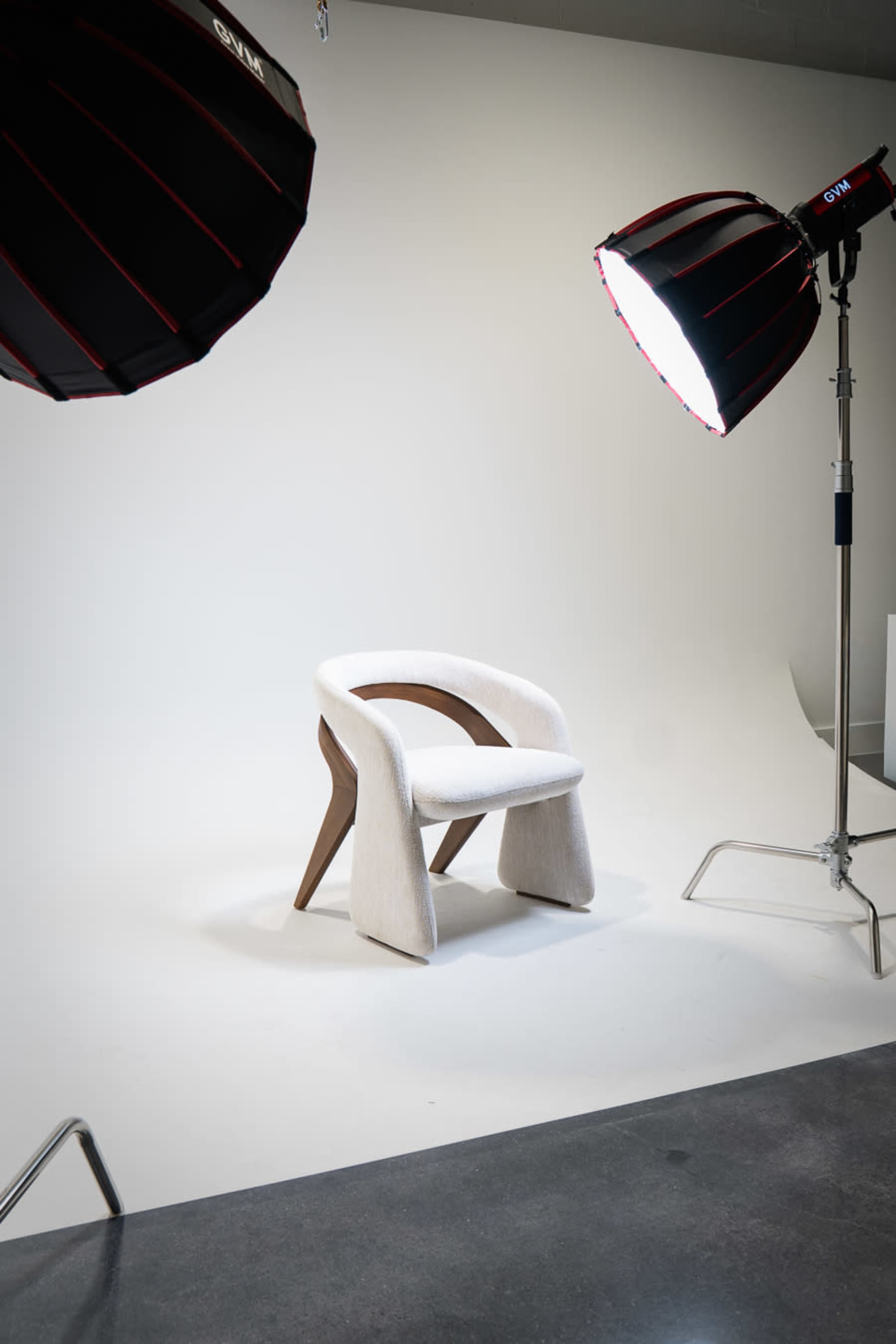 A soft beige chair with wooden legs is positioned in an empty studio space illuminated by two large softbox lights.
